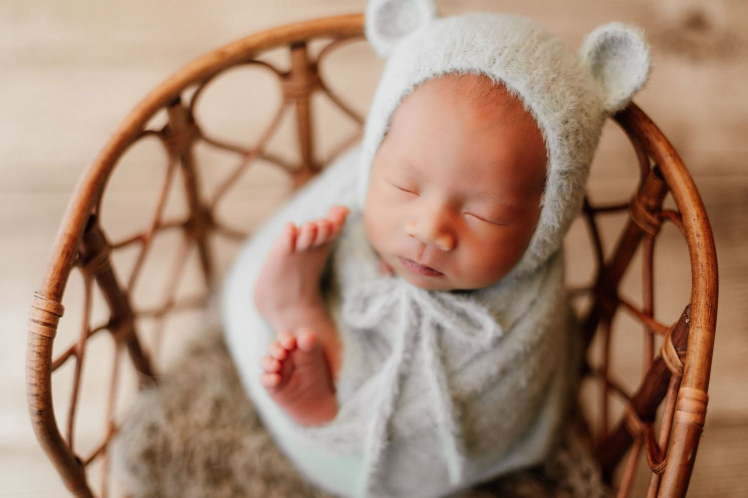 Peaceful newborn baby sleeping in a wicker chair, wrapped in gray knit, wearing a bear ear bonnet.