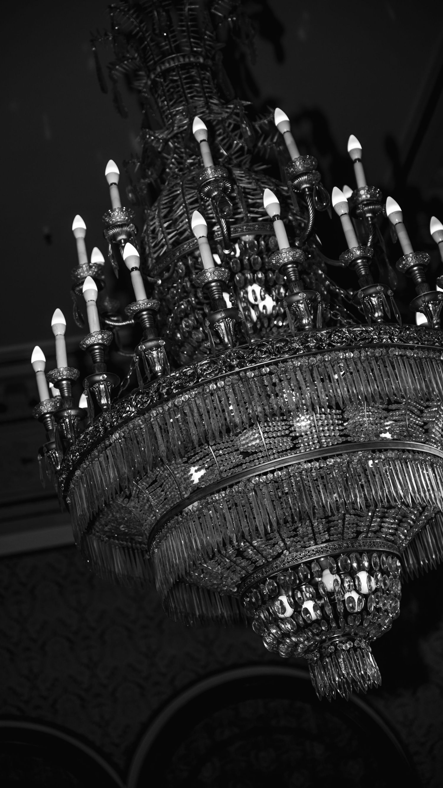 A dramatic black and white image of an ornate crystal chandelier with multiple tiers of candle-style lights.