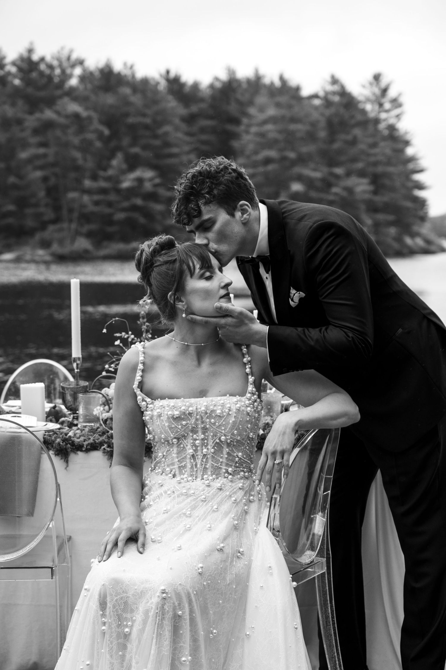 A romantic black and white wedding portrait captures an intimate moment between newlyweds near a scenic lake with pine trees.