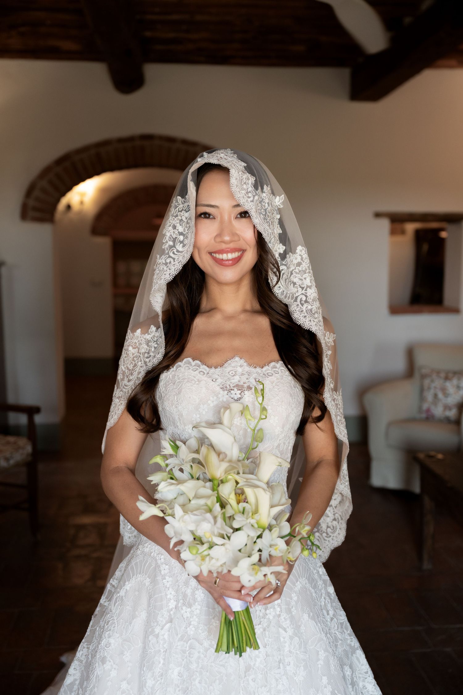Smiling Asian bride in lace wedding gown and veil holds white calla lily bouquet indoors.