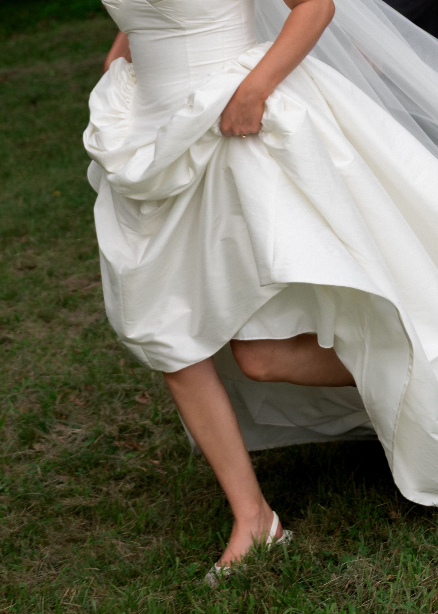Bride in a white wedding gown lifting her dress hem, revealing flat bridal shoes while standing on green grass.