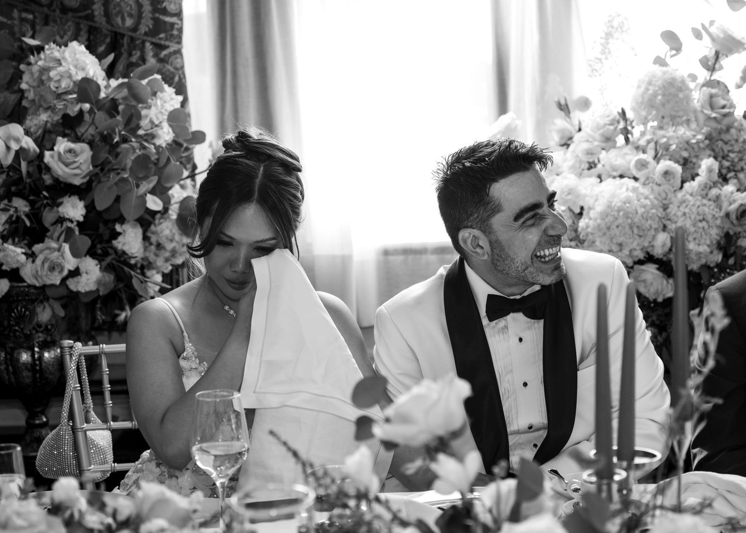 A black and white photo shows a couple seated at an elegantly decorated wedding reception table with floral arrangements.