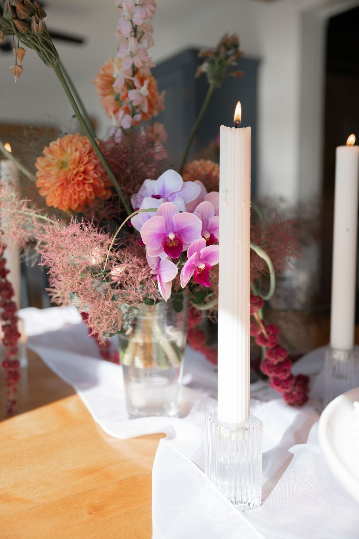 A lit white candle beside a floral arrangement with pink orchids, orange dahlias and soft pink astilbe in a glass vase.