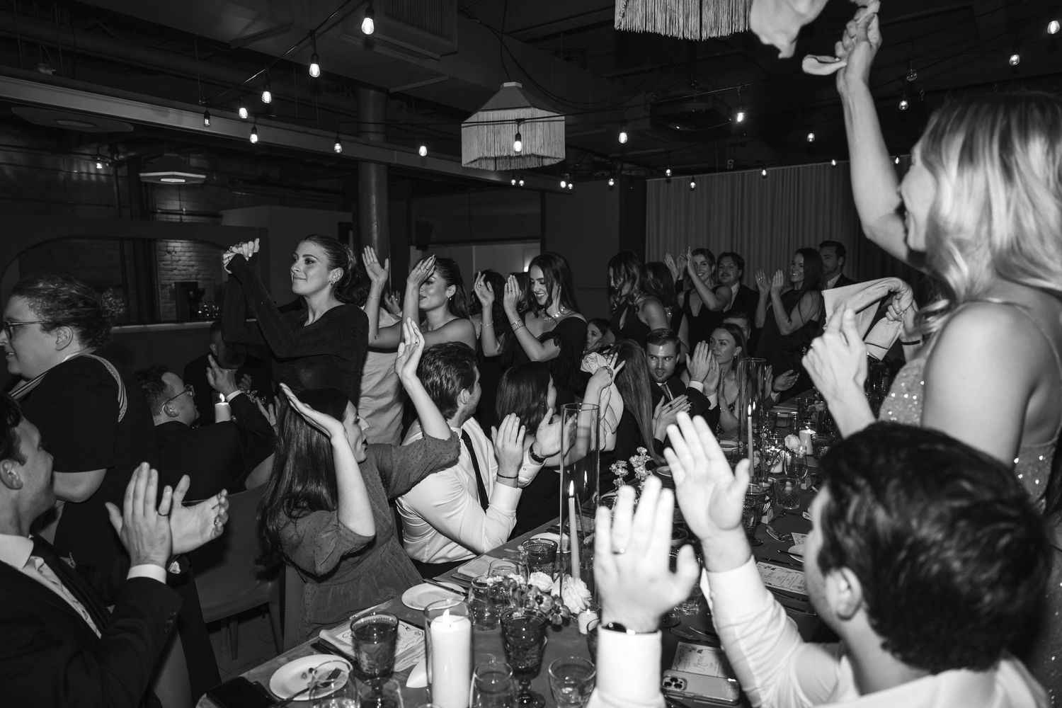 A crowded nightclub with people dancing and raising their hands in a celebratory atmosphere captured in black and white.