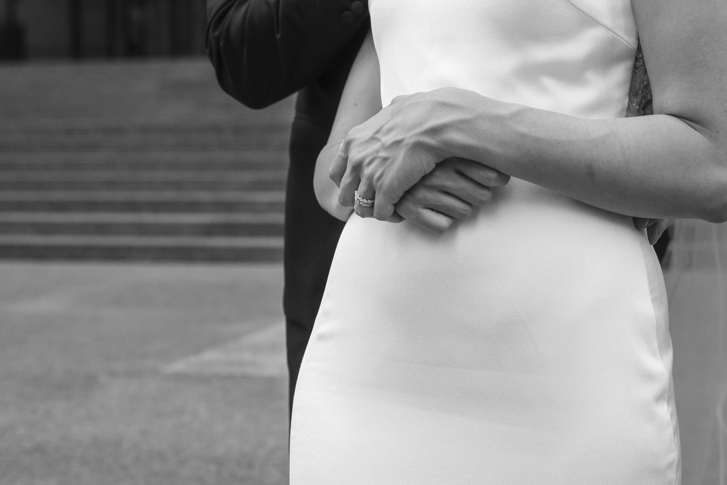 Black and white close up of couple embracing, showing hands gently resting on a white dress.