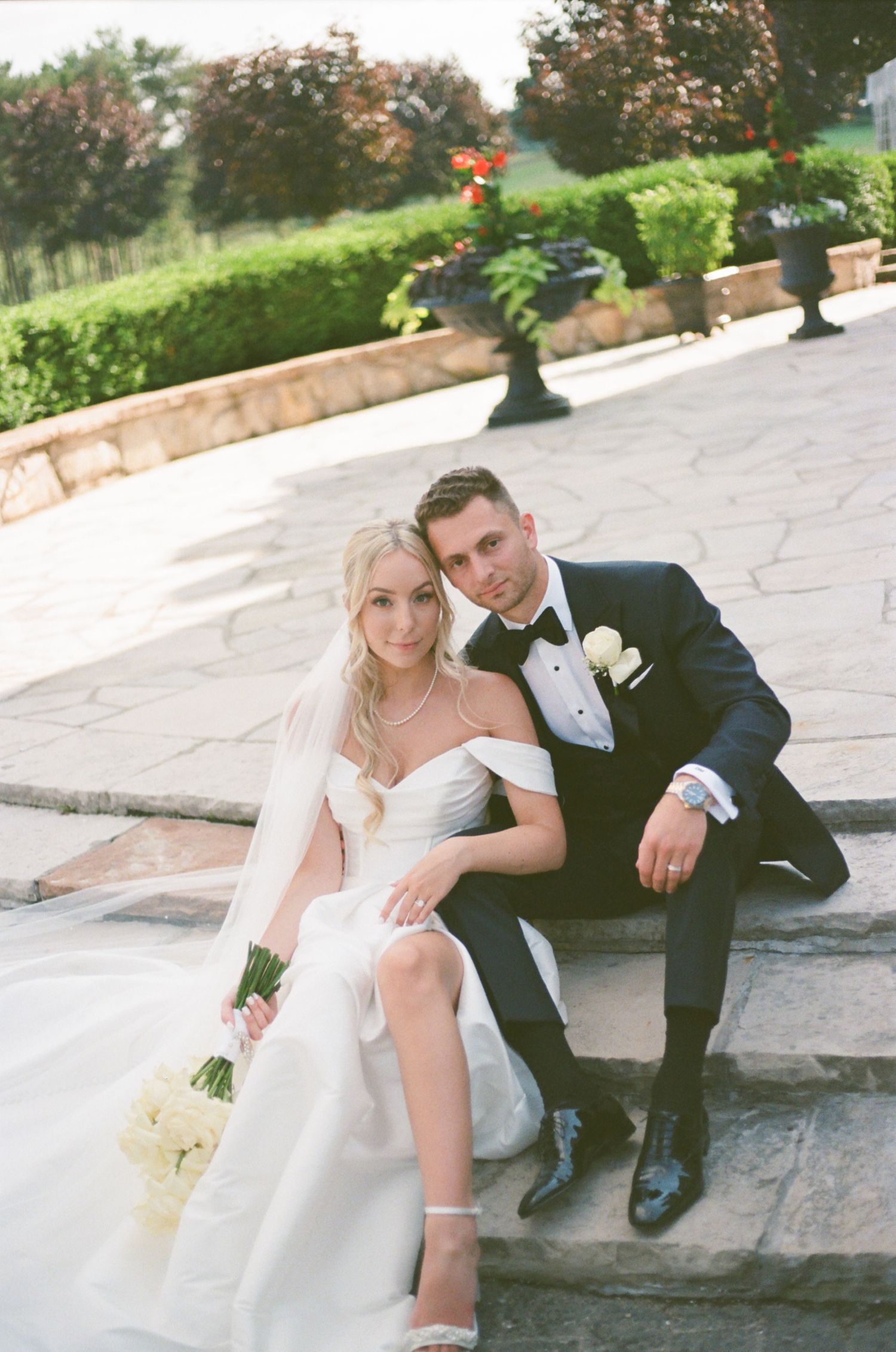 A romantic wedding couple sits together on stone steps surrounded by lush greenery and gardens.