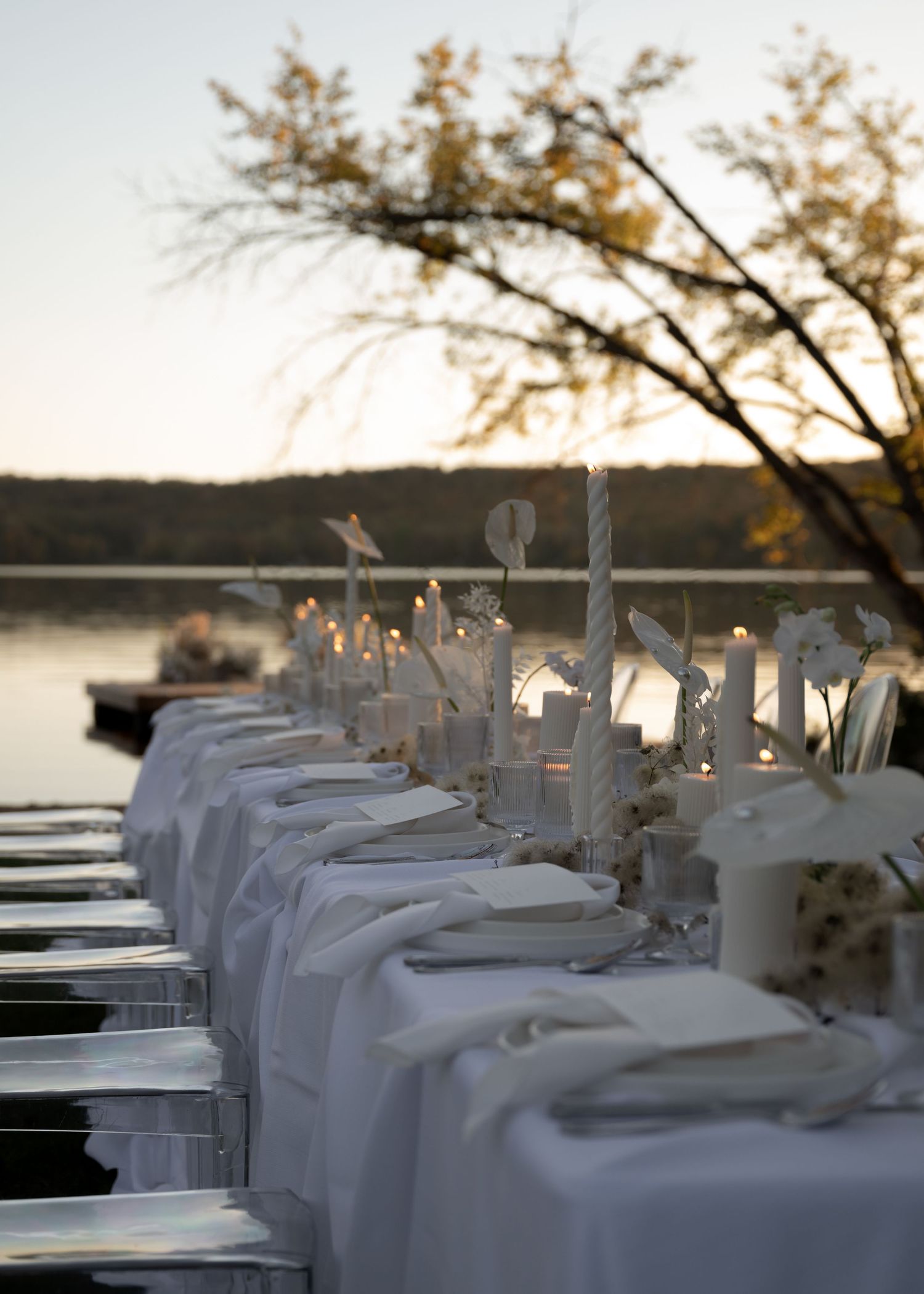 Elegant outdoor dining table set with white linens and glowing candles beside a peaceful lake at sunset.