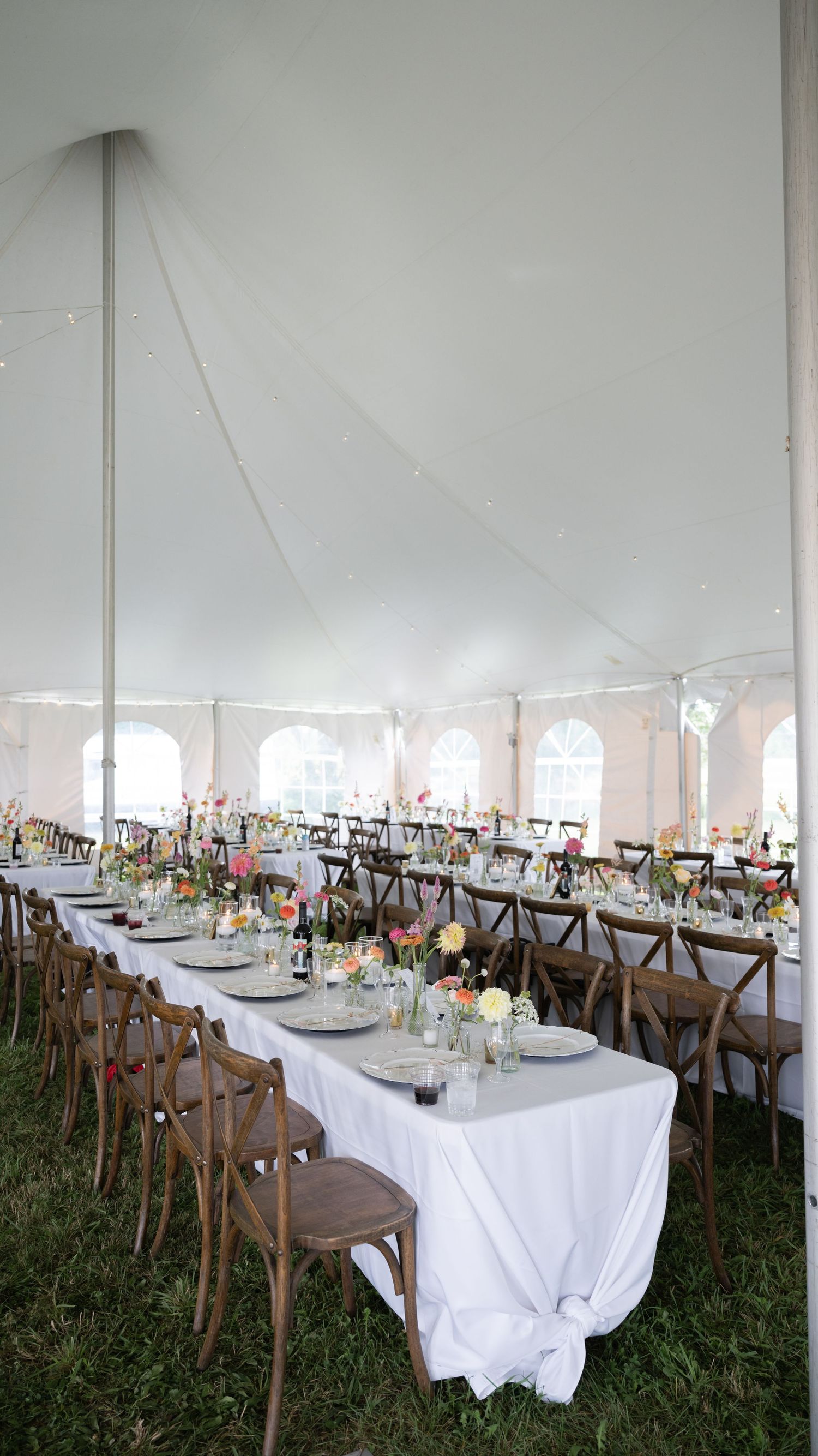 Elegant wedding reception setup under a white tent with long tables and cross-back chairs.