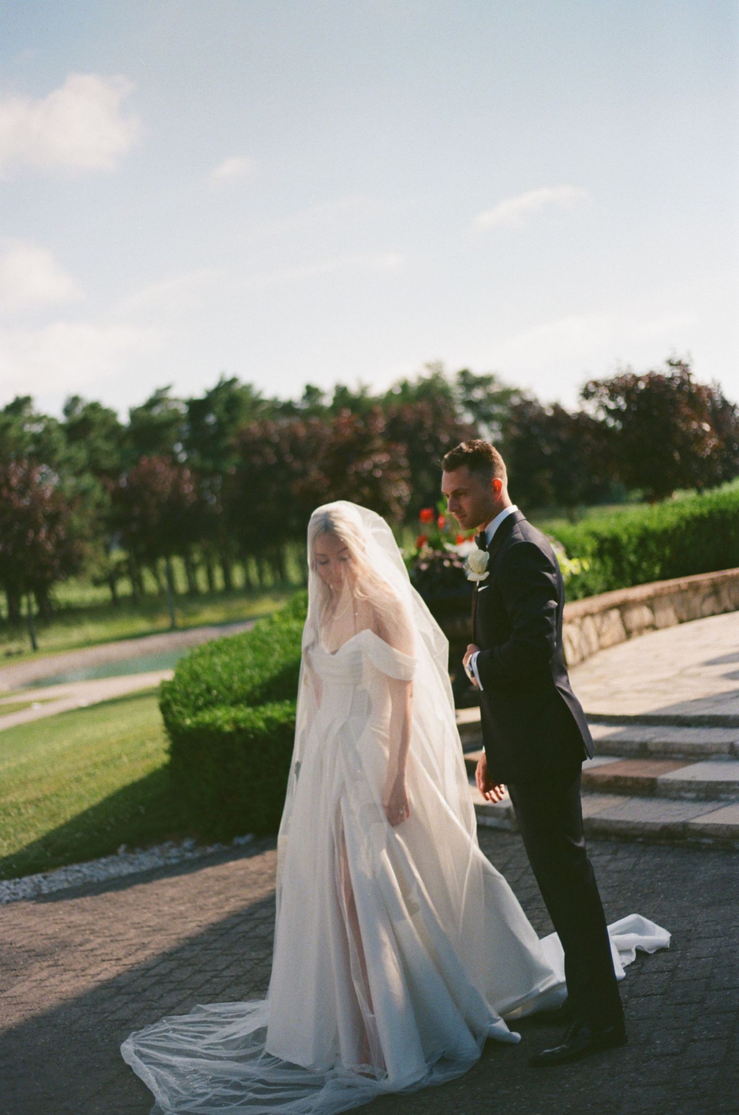 A couple in wedding attire stand on a curved path with greenery and trees in the background during sunset.