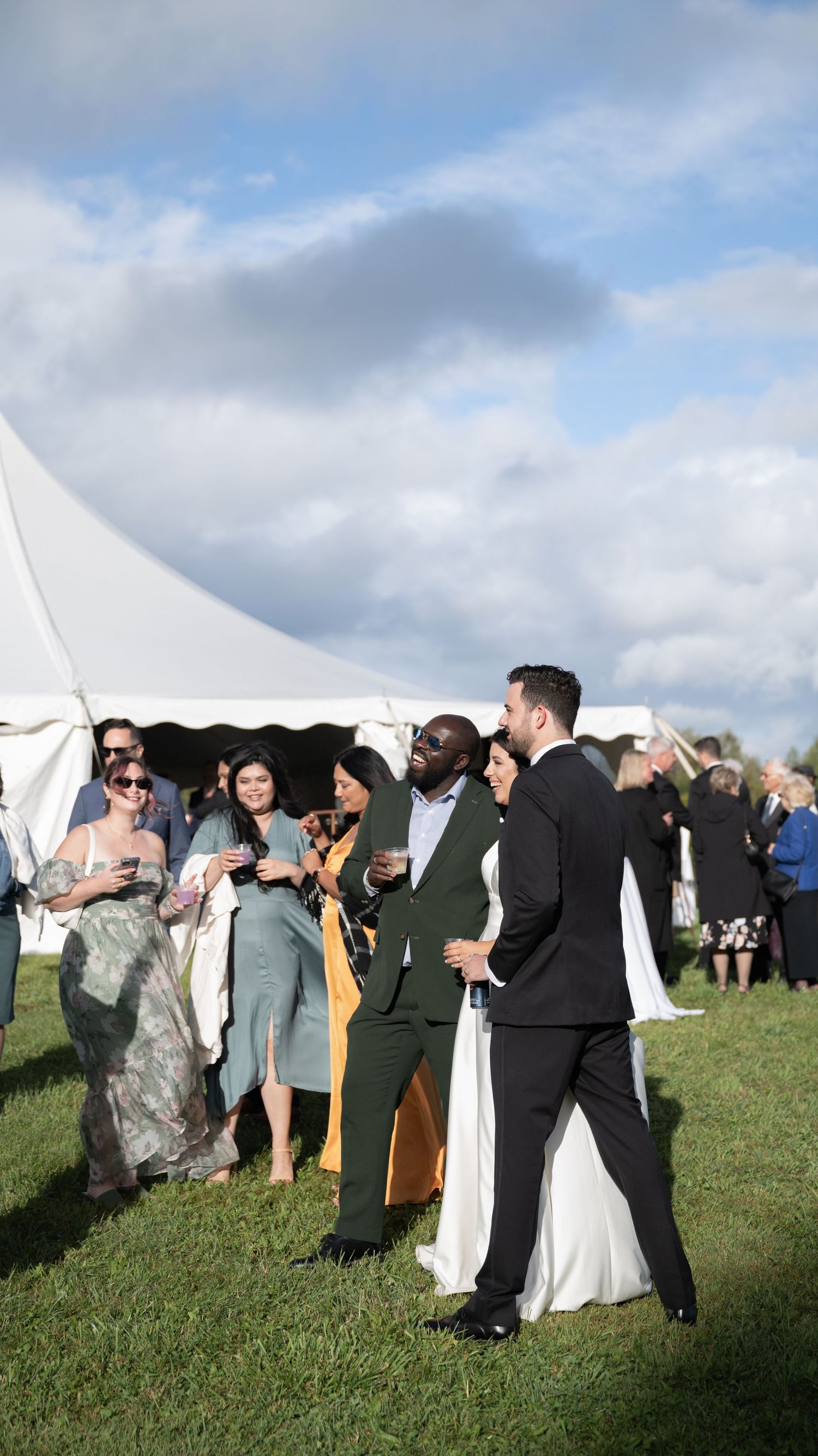 Wedding guests mingle outdoors near white event tents on a grassy lawn under cloudy skies.