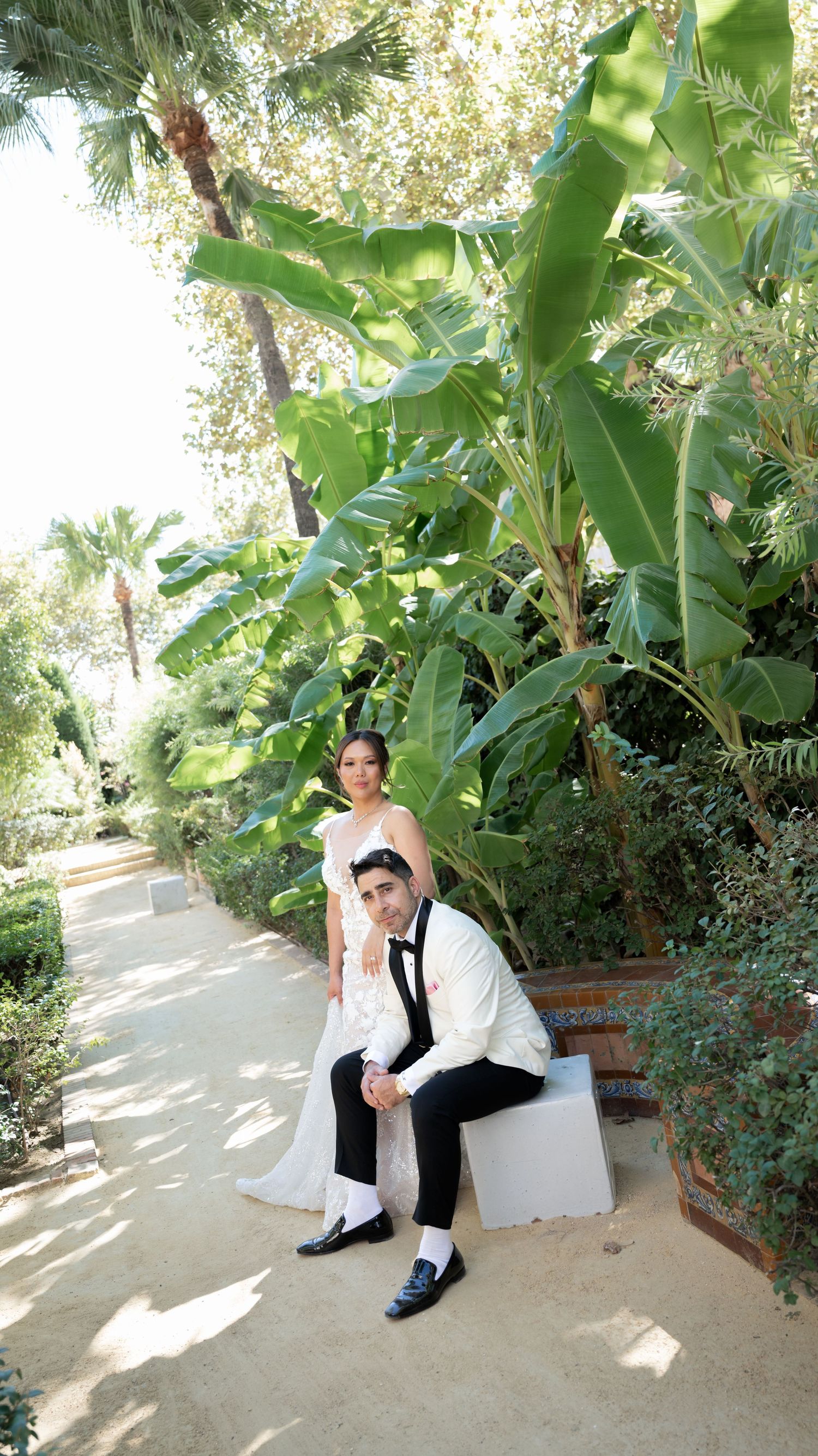 A bride and groom pose together on a garden path surrounded by lush tropical plants and palm trees.