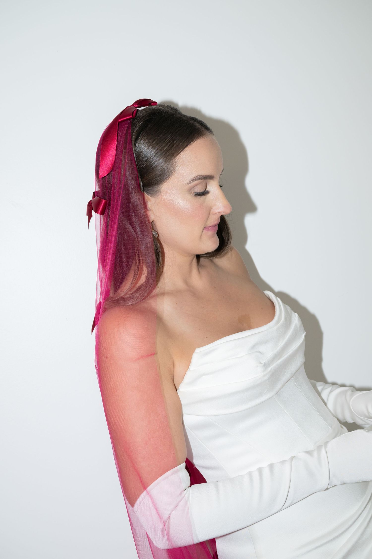 Woman in white formal attire with pink hair ribbon poses against light backdrop in elegant portrait.