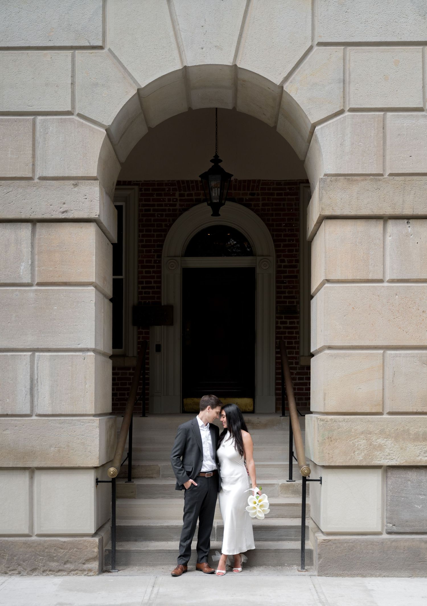 A couple shares a romantic moment under a grand stone archway entrance with elegant architecture and an iron lantern above.