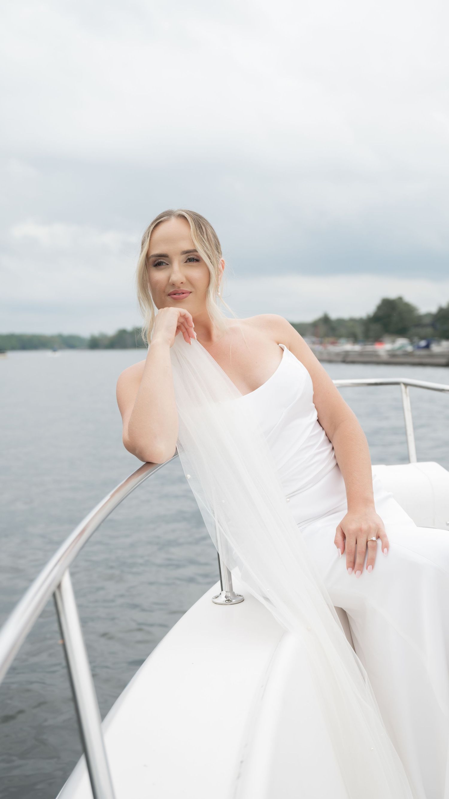 A bride in an elegant white wedding dress poses on a boat deck with water and cloudy sky in the background.