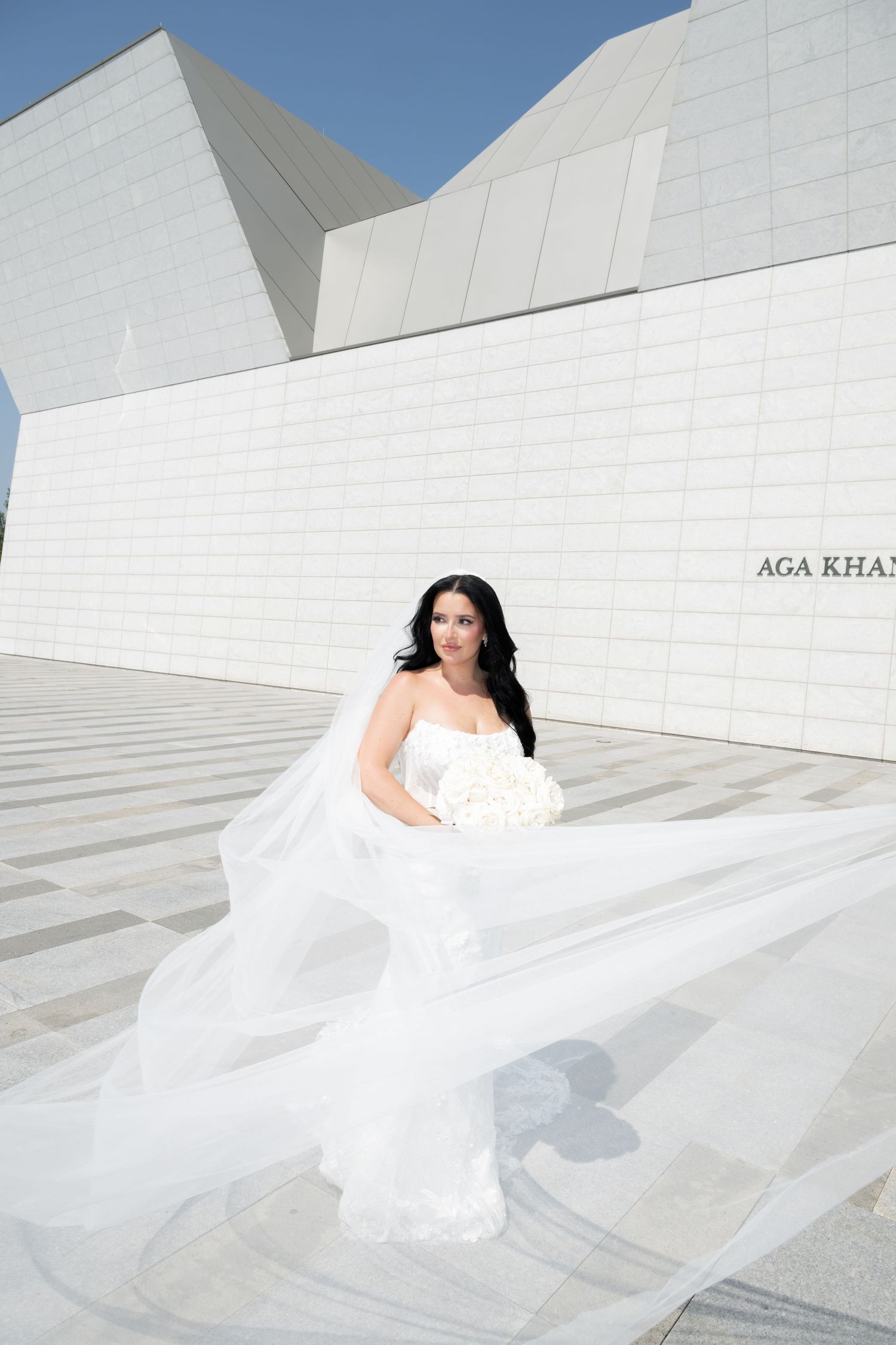 A bride in a flowing white gown poses on sleek modern white architectural stairs under bright blue sky.