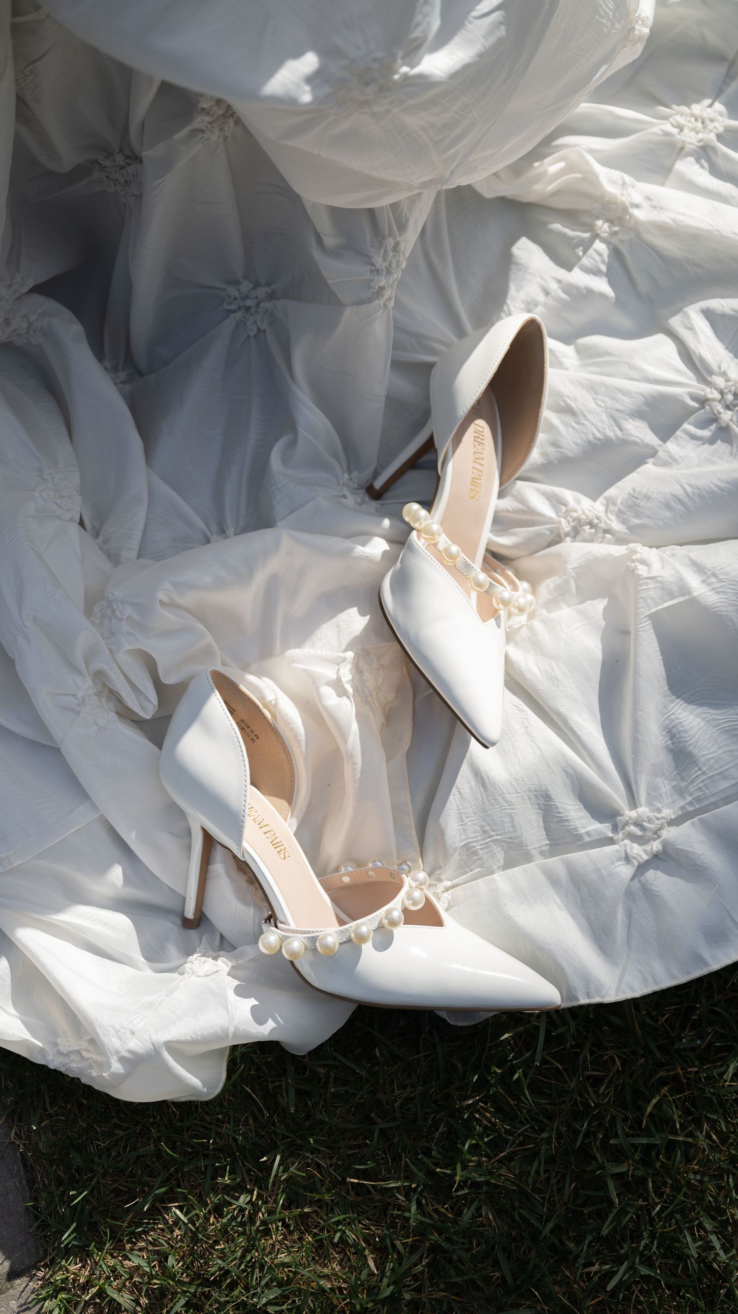 Elegant white bridal heels with bow details resting on flowing fabric outdoors.