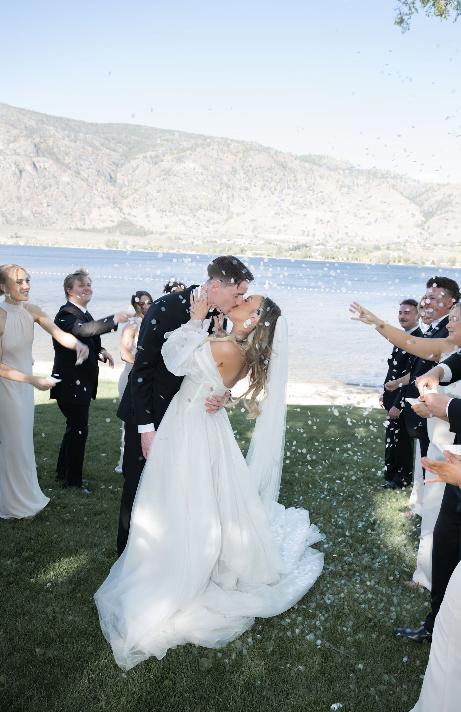 A romantic outdoor wedding ceremony by a lake with mountains in the background as guests shower the couple with flower petals.