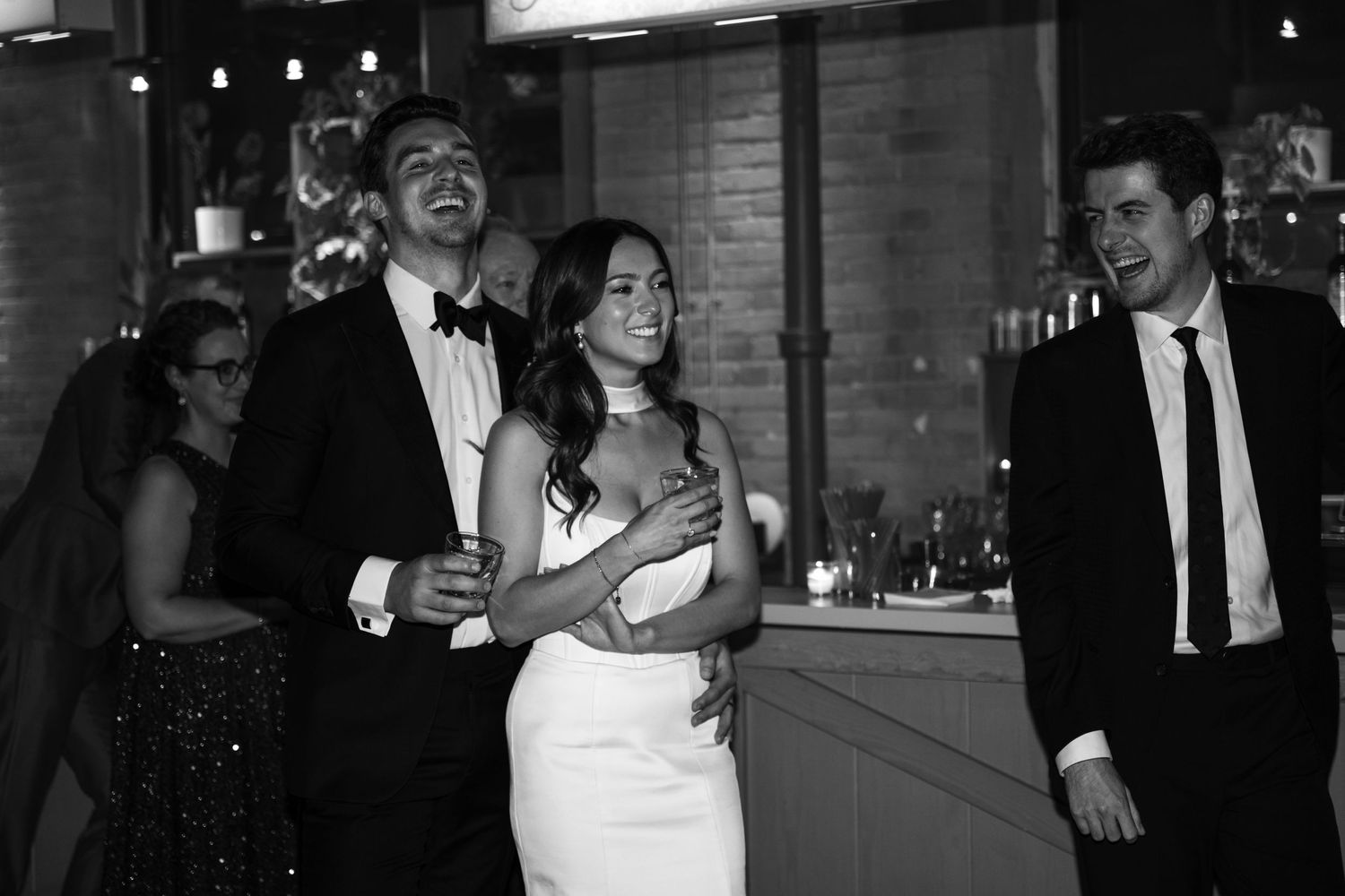 Black and white candid photo showing wedding reception celebration with guests laughing together.