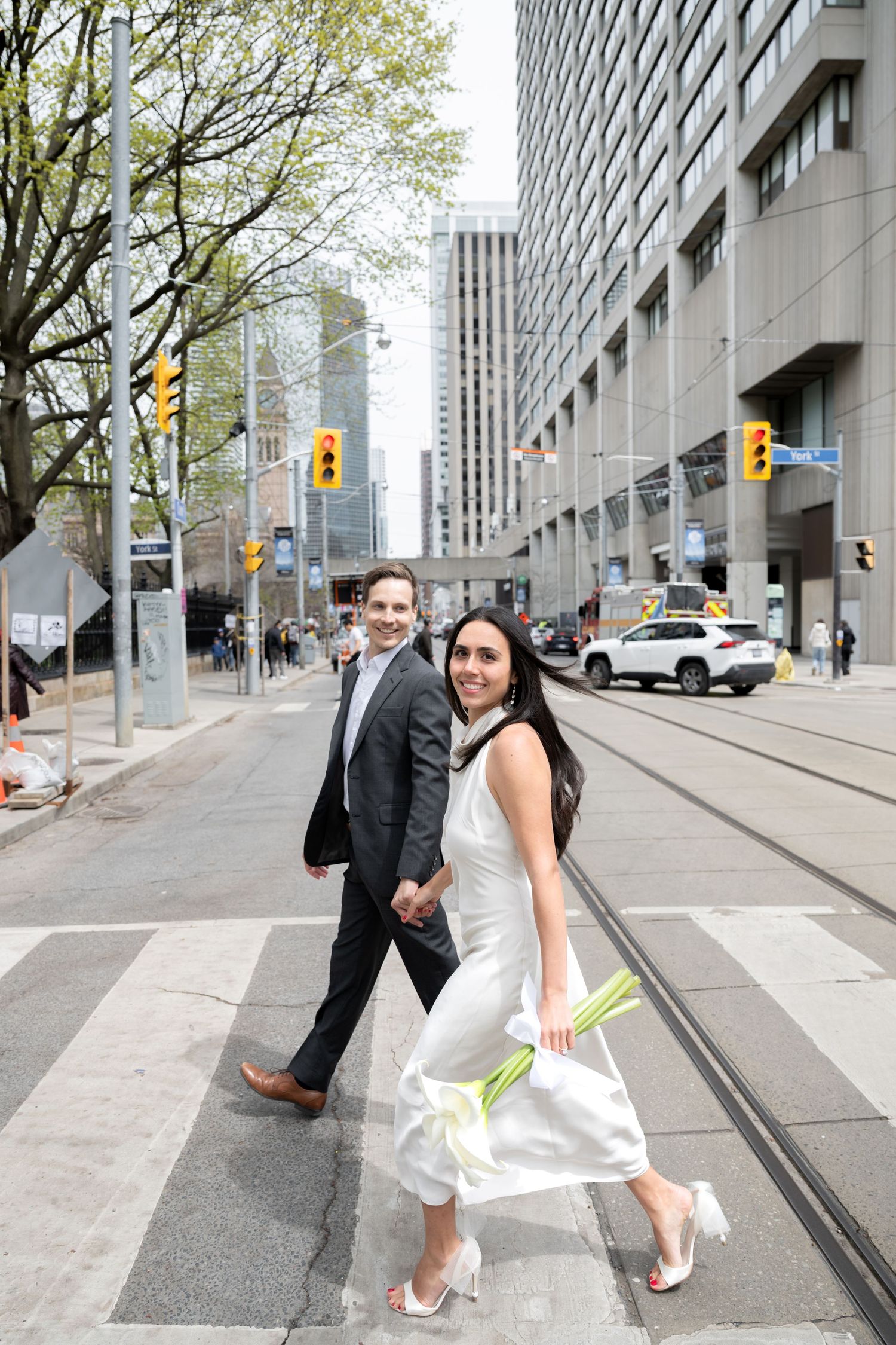 A couple in formal attire walking across a downtown intersection near streetcar tracks on a city street.