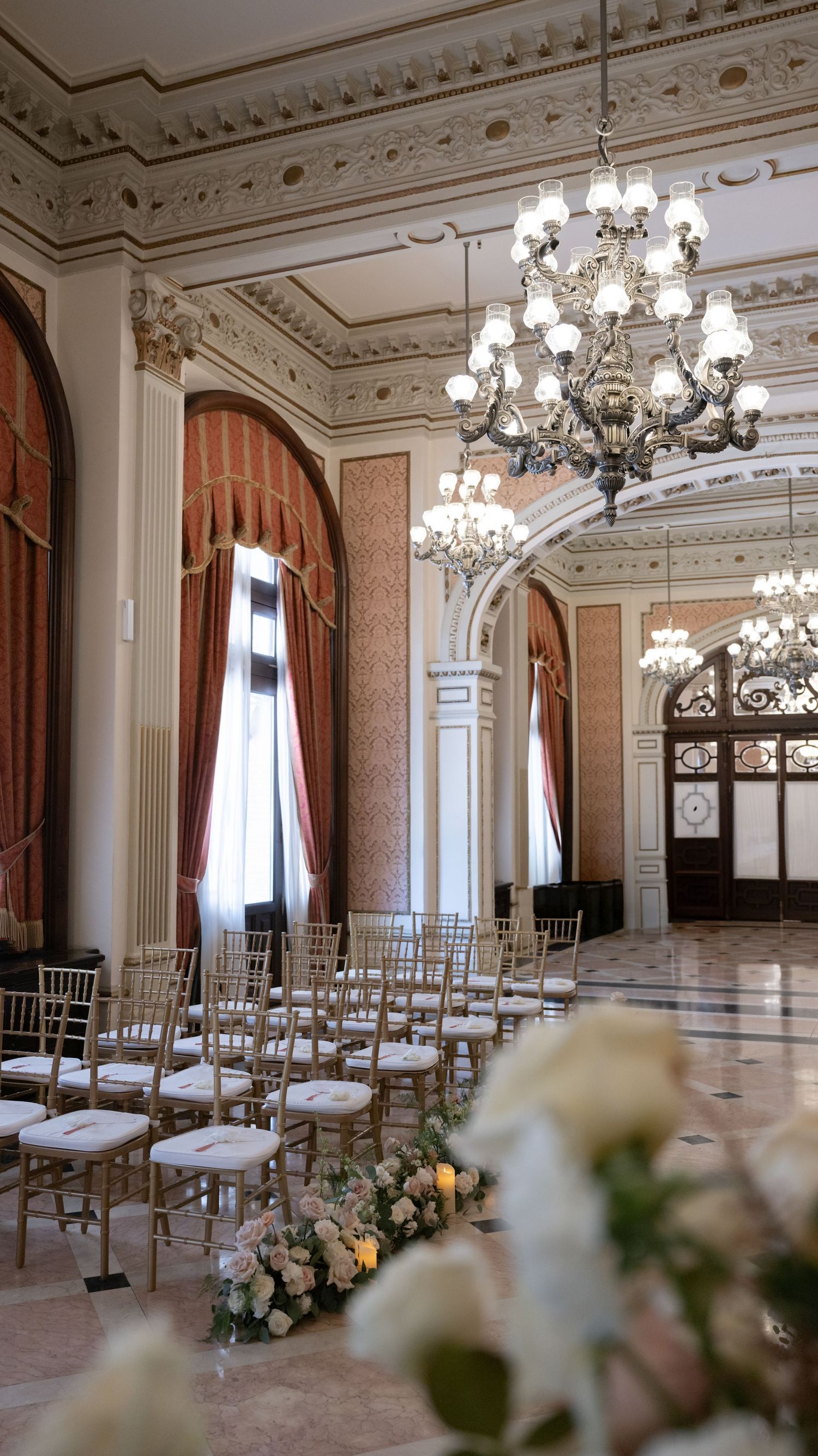 Elegant ballroom with ornate chandeliers and chairs set up for a wedding ceremony with floral decorations.