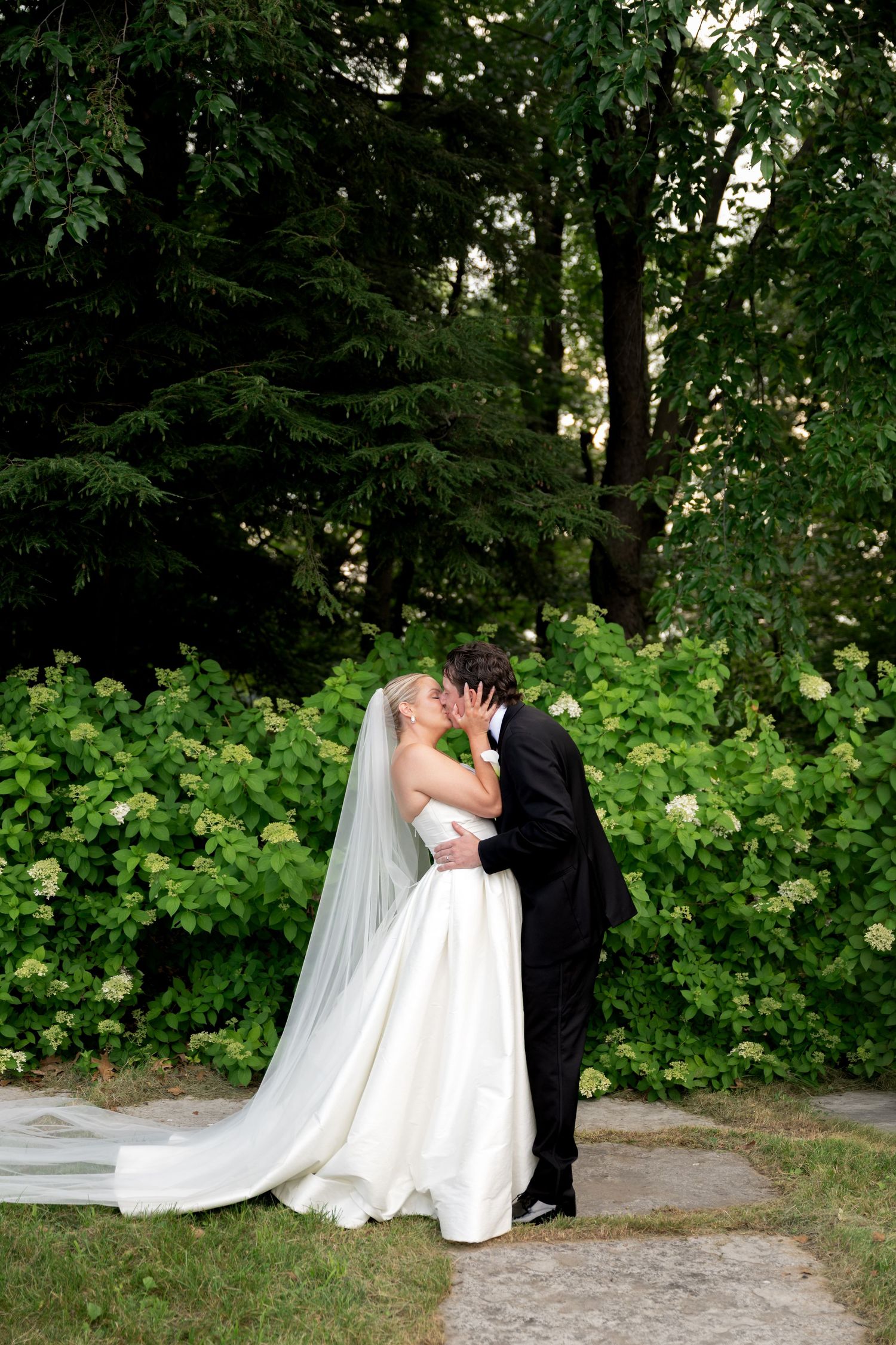 Bride and groom share a romantic kiss outdoors, surrounded by lush green trees and white hydrangeas.