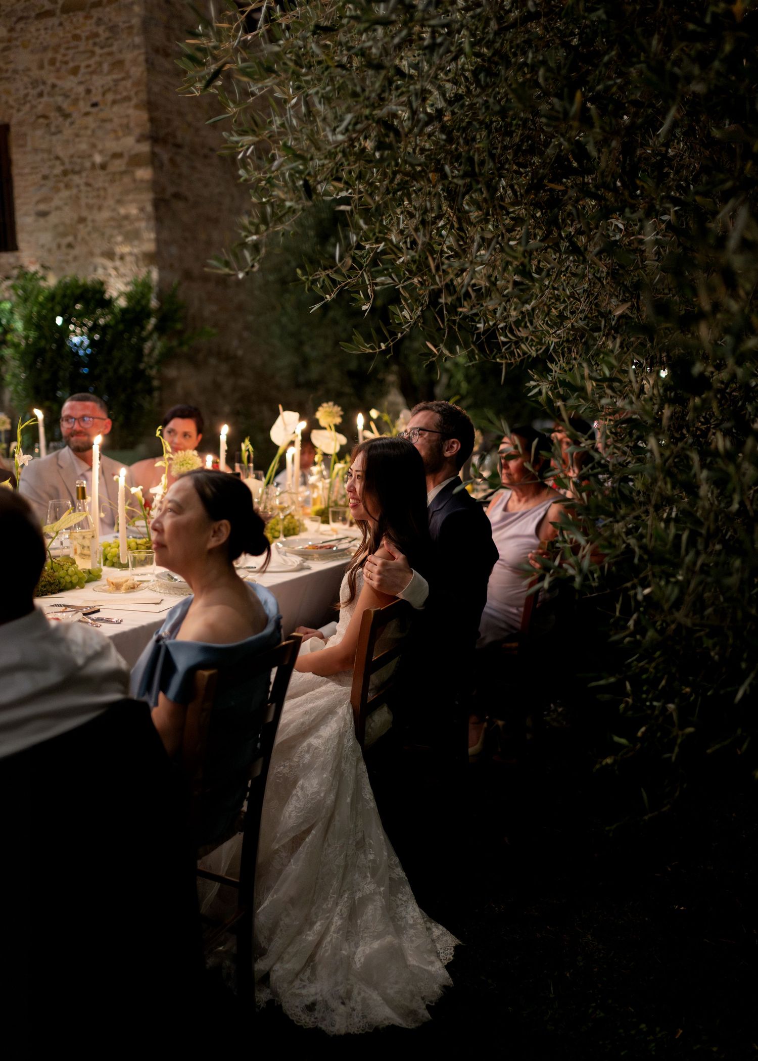 Bride and groom share a romantic outdoor wedding dance at night, surrounded by candlelit tables and olive trees.