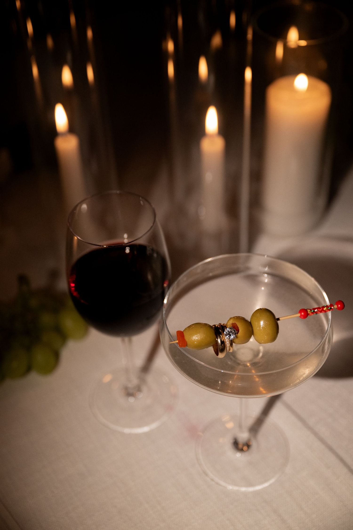 Elegant martini with olive garnish and red wine glass by candlelight in romantic setting.