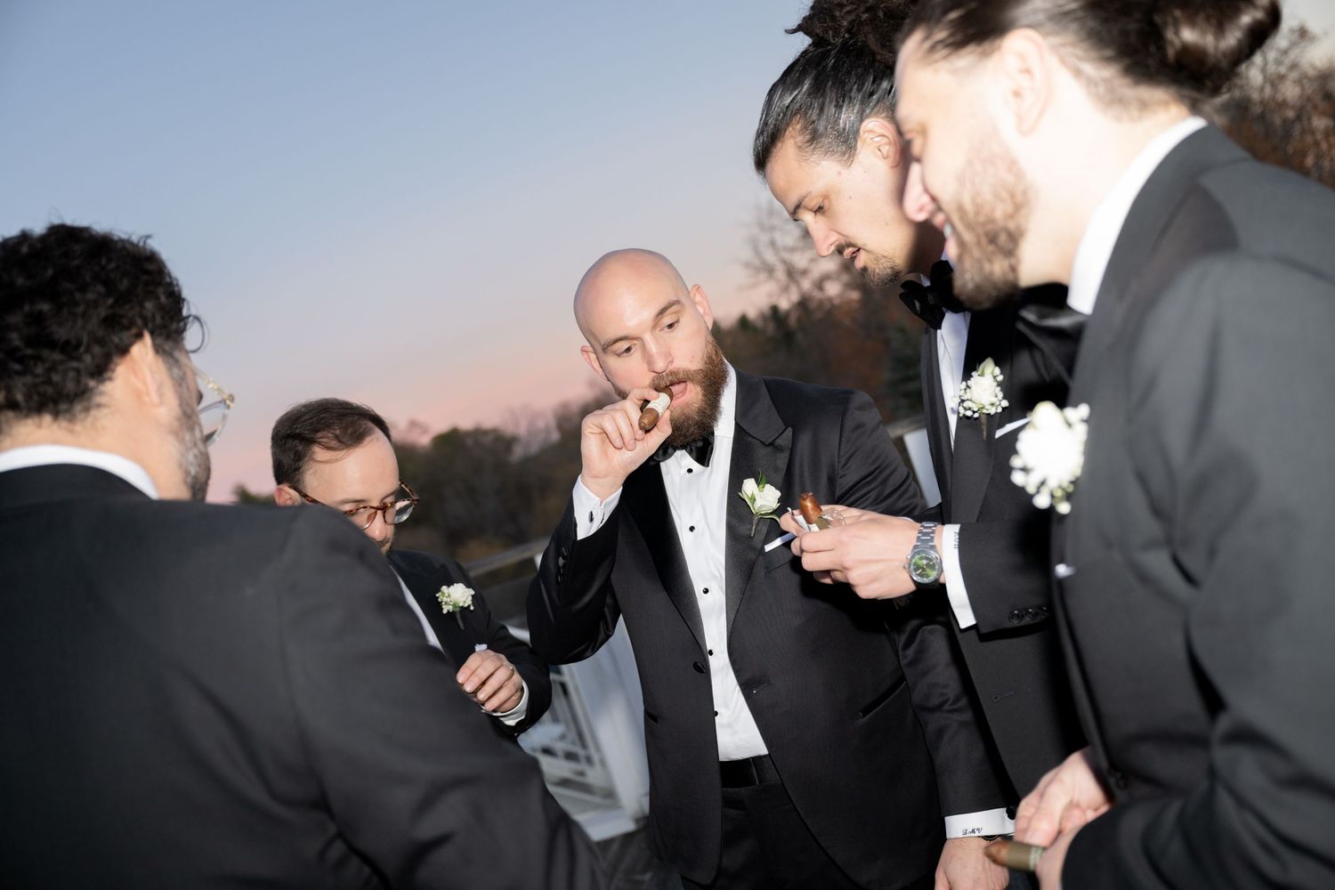 A group of men in formal attire laugh and share a moment together during an outdoor evening celebration.