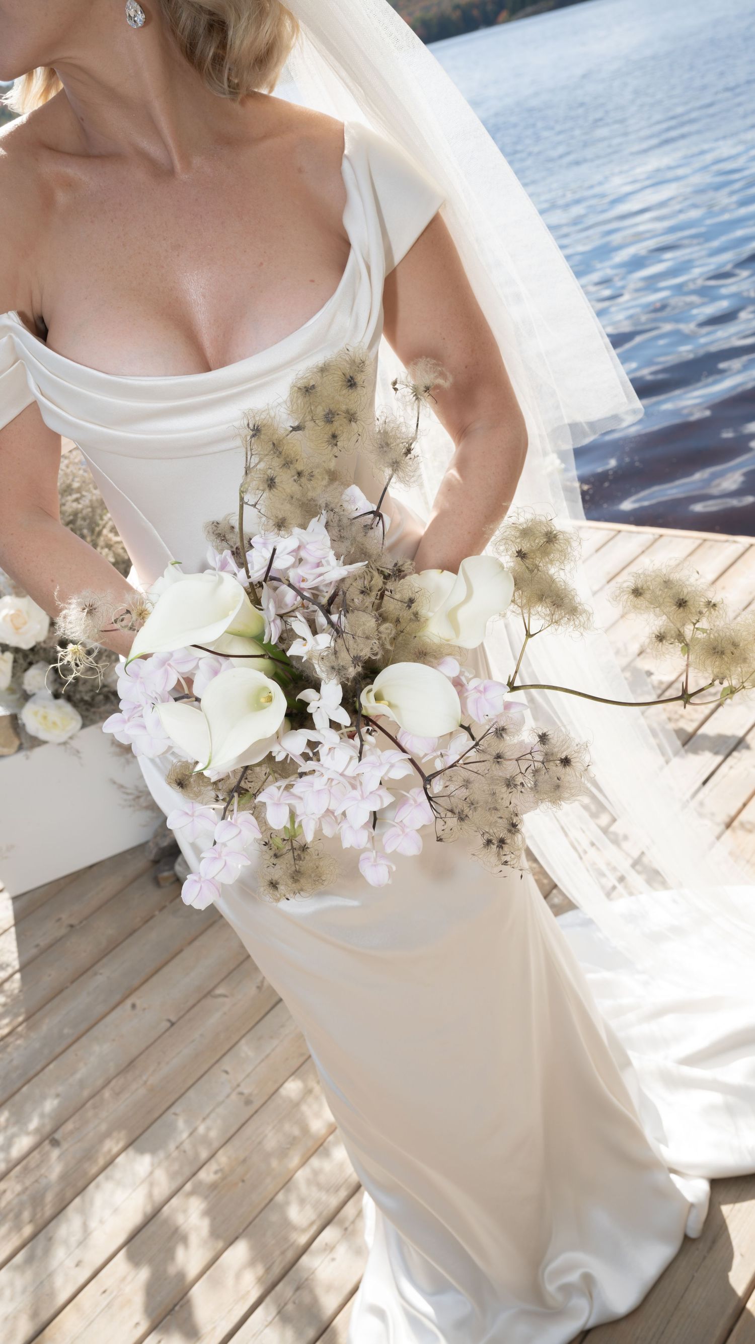 A stunning white wedding dress and delicate floral bouquet of white roses and baby's breath on a wooden dock by the water.