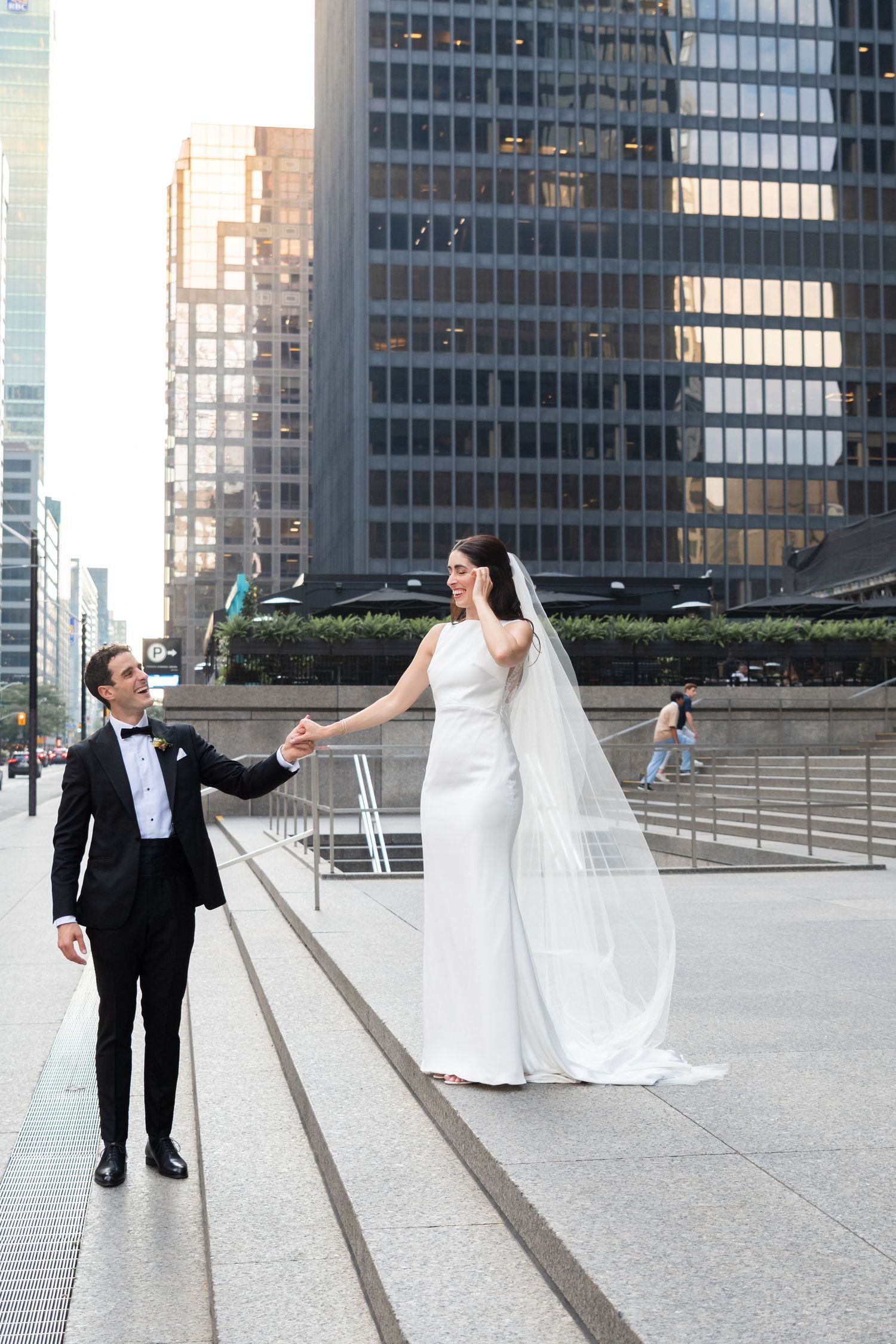 Elegant wedding couple holding hands on urban walkway with modern skyscrapers in background.