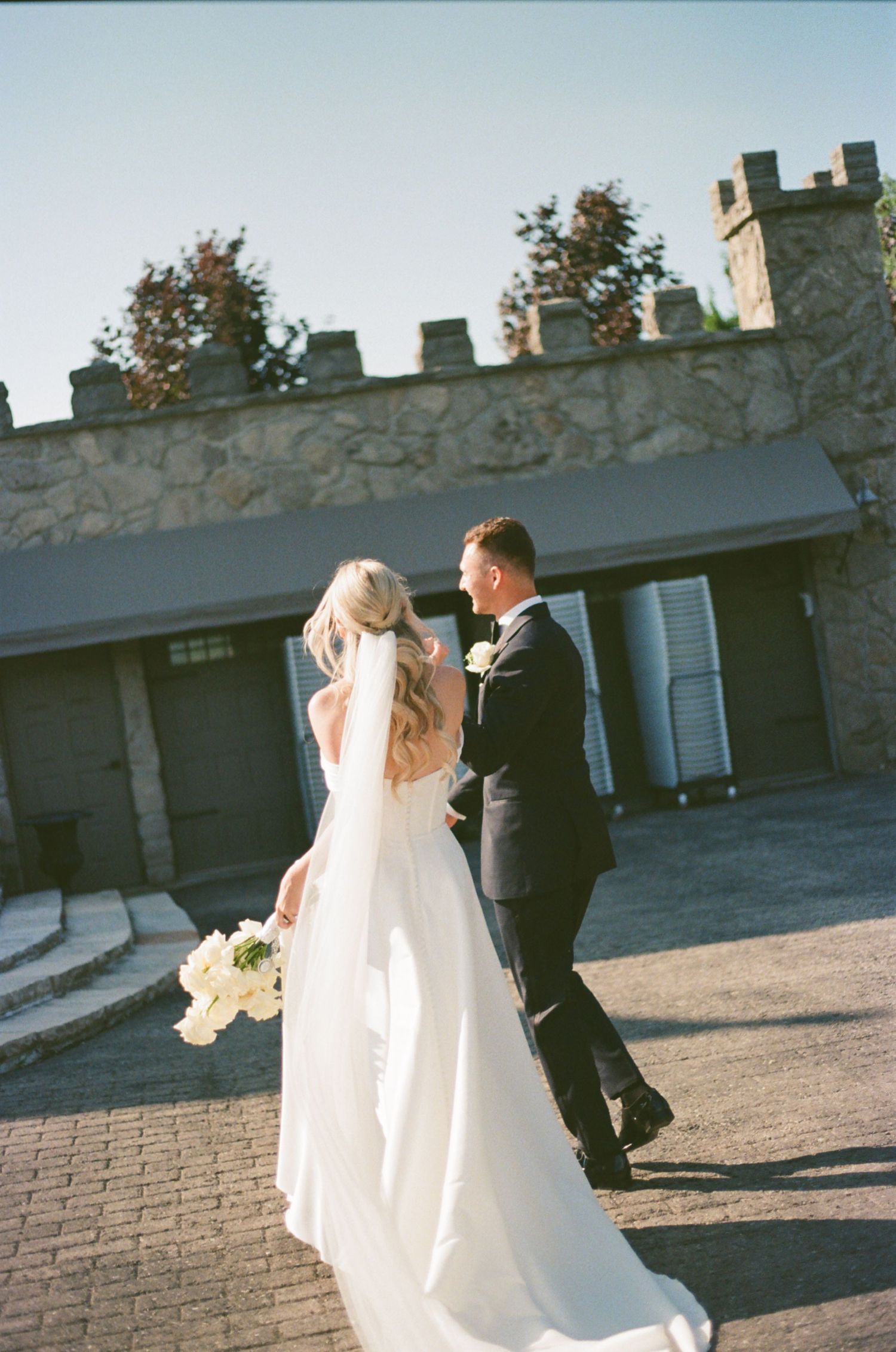 A couple in wedding attire share a moment together on a brick patio with stone walls in the background.