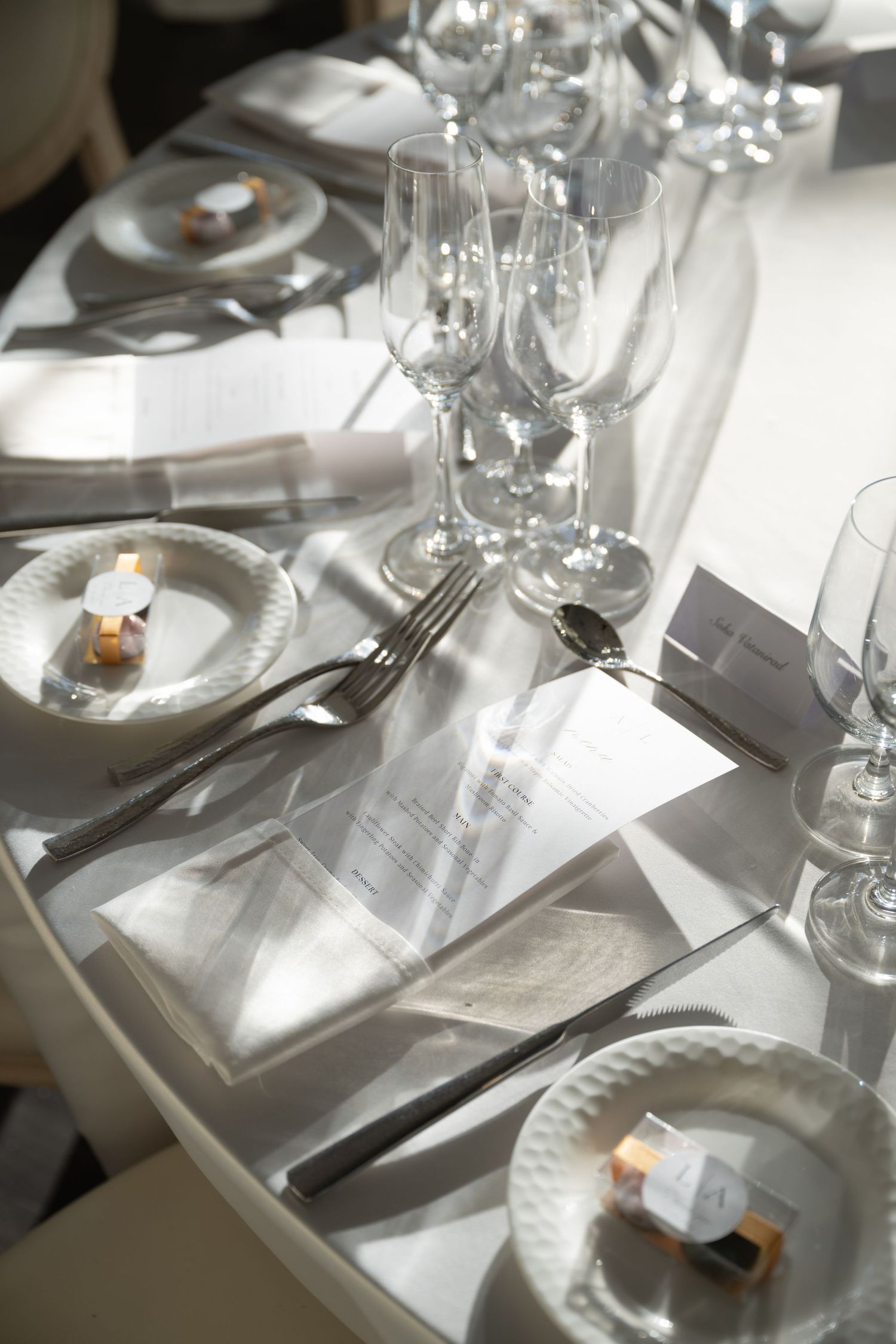 Elegant white table setting with wine glasses, plates, and place cards in natural sunlight creating dramatic shadows.