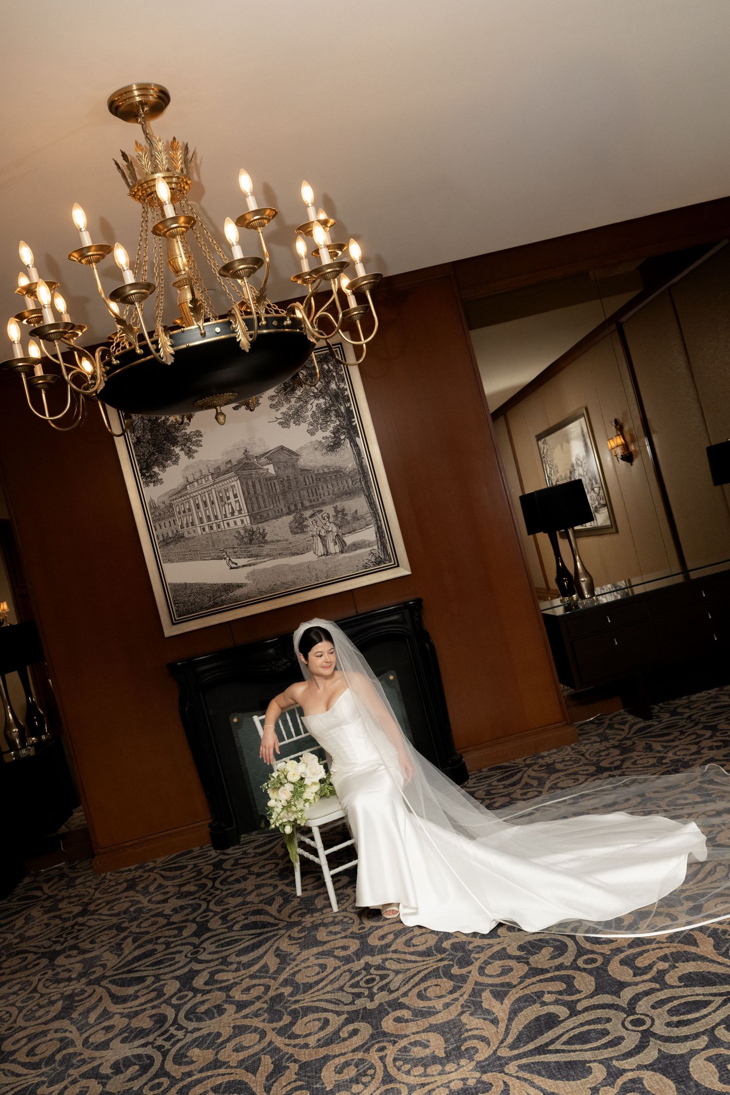 Elegant bride in white gown and veil seated by fireplace under gold chandelier in luxurious wedding venue.