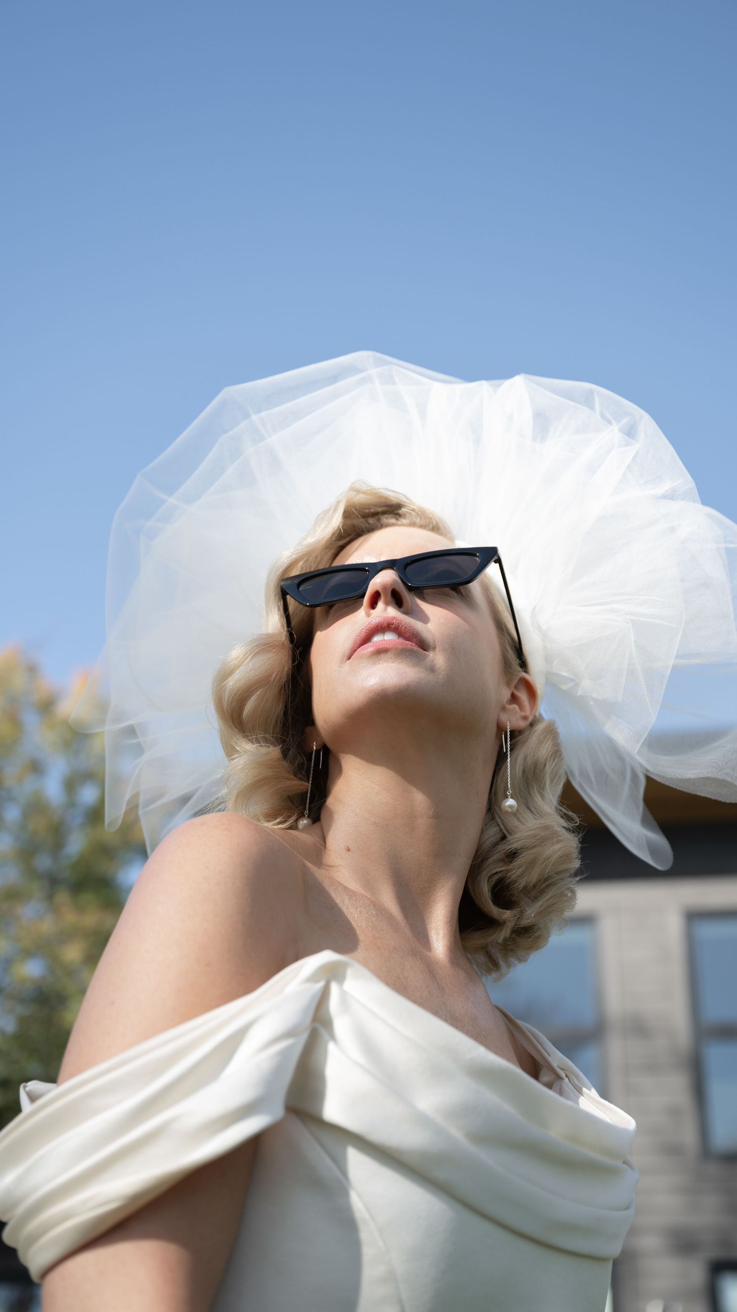 Stylish bride in sunglasses and off-shoulder dress against blue sky.