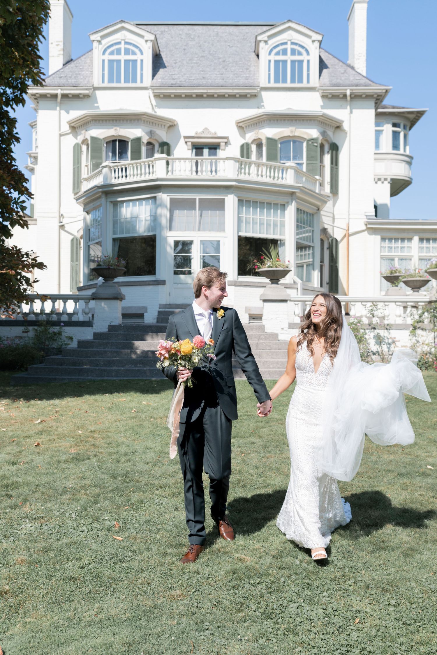 Newlywed couple runs joyfully across the lawn of a grand white mansion with dormer windows and elegant architecture.