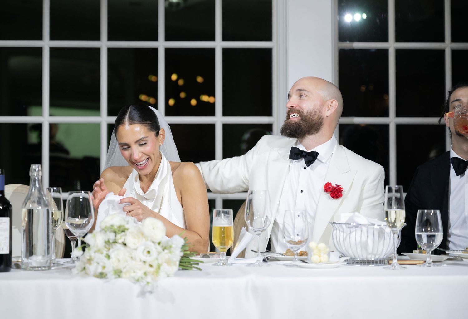 A couple shares a candid moment at their wedding reception head table with elegant place settings and floral decor.