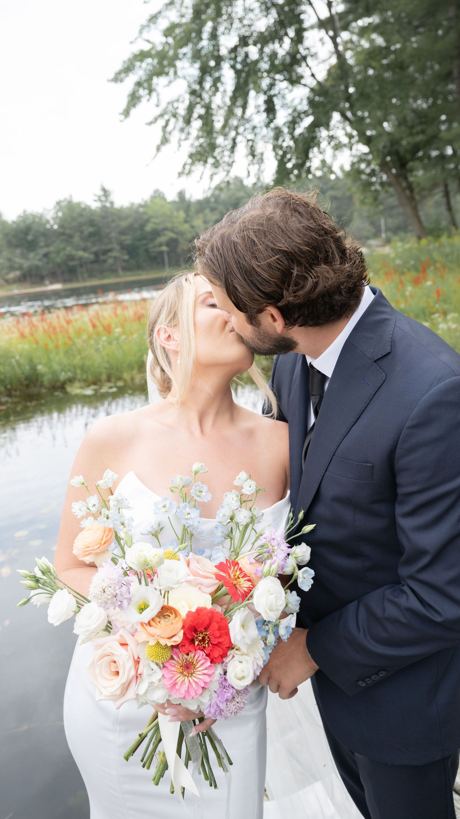 A newlywed couple shares a kiss by a pond while the bride holds a vibrant bouquet of pink, red and white flowers.