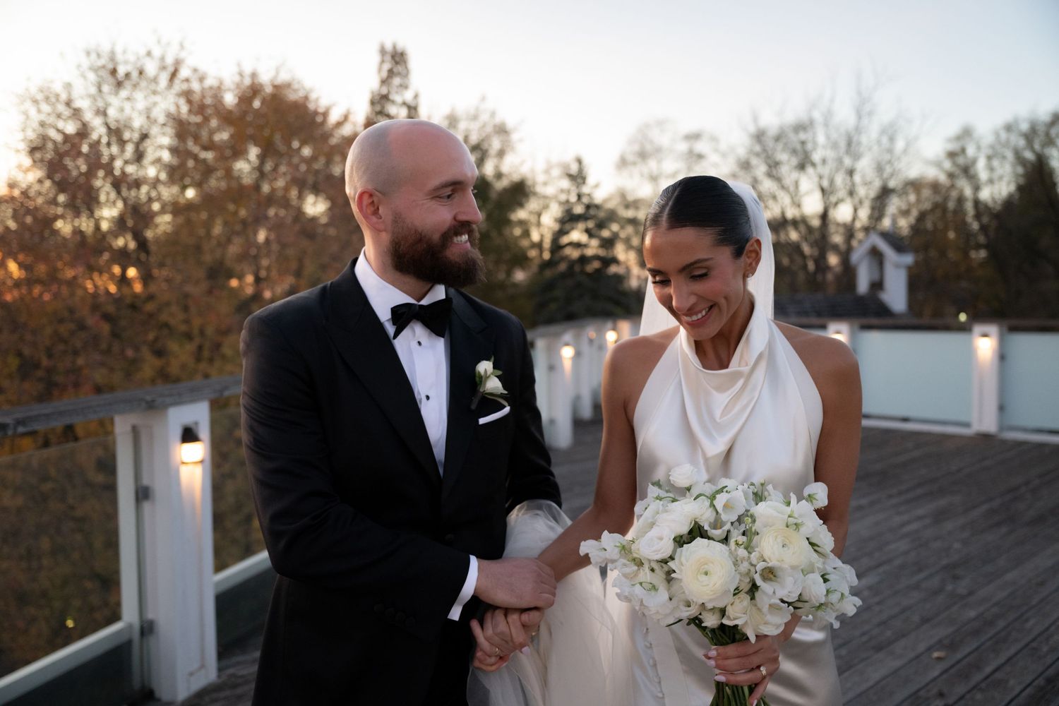 A newlywed couple shares a joyous moment together on a deck during golden hour with a white floral bouquet.