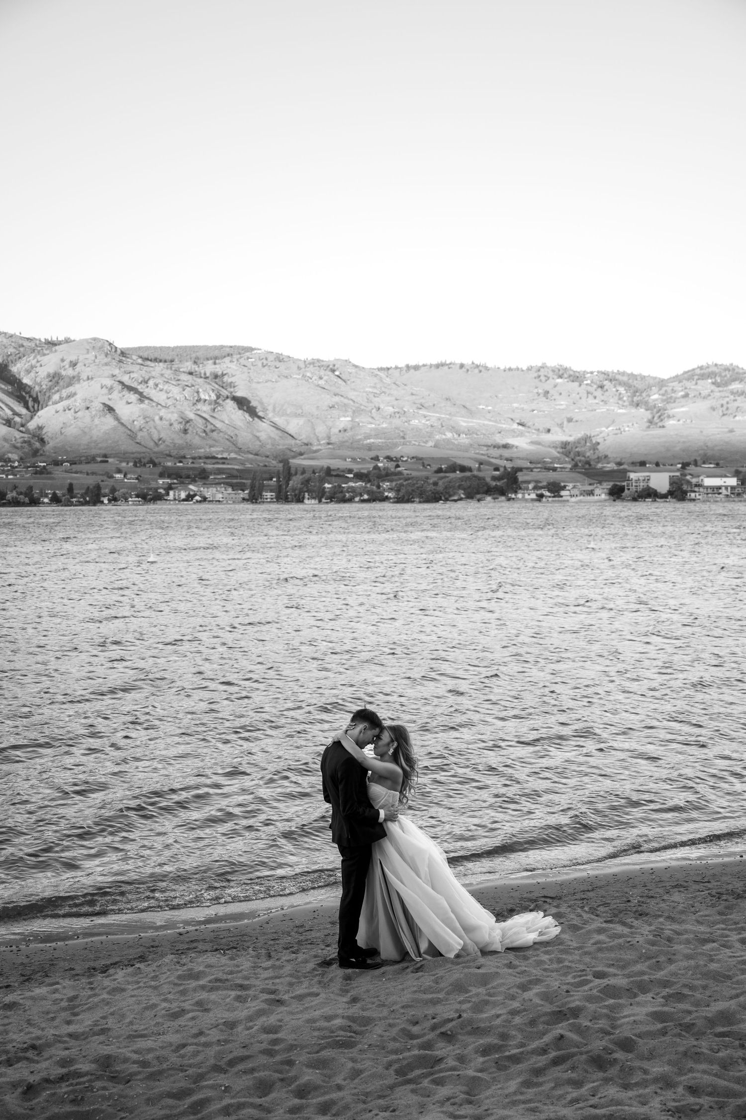 A couple embraces on a beach shore with mountains and coastline in the background, captured in black and white.