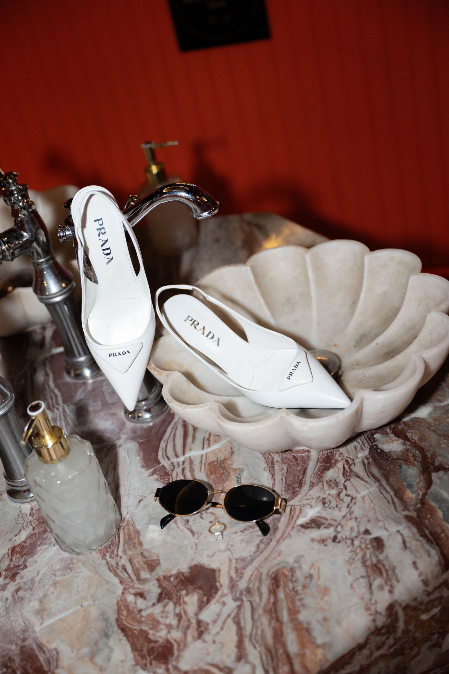 White designer heels and sunglasses displayed on a marble surface against a red wall.
