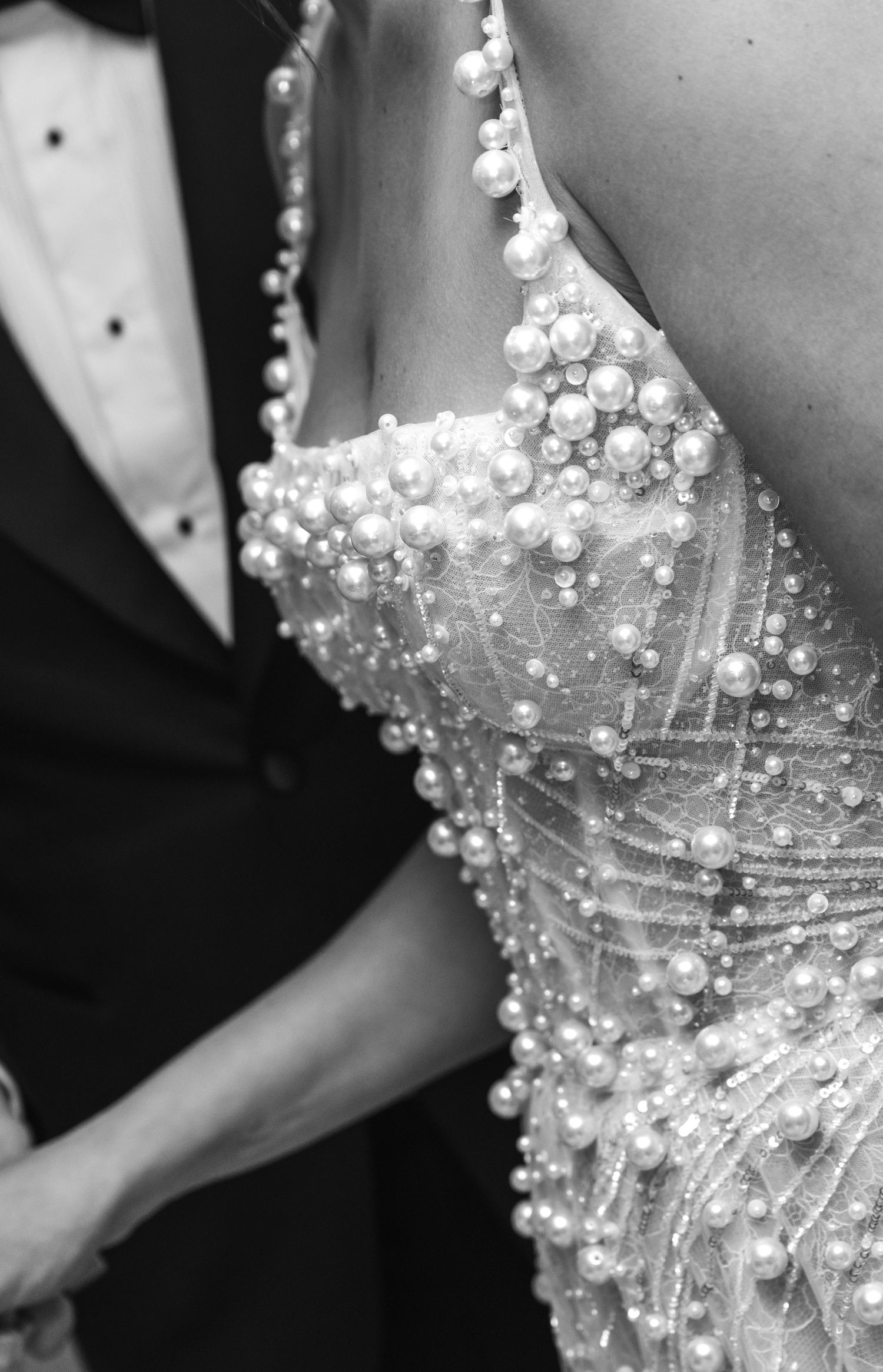 Close-up detail of a pearl-embellished wedding dress beside a black tuxedo.