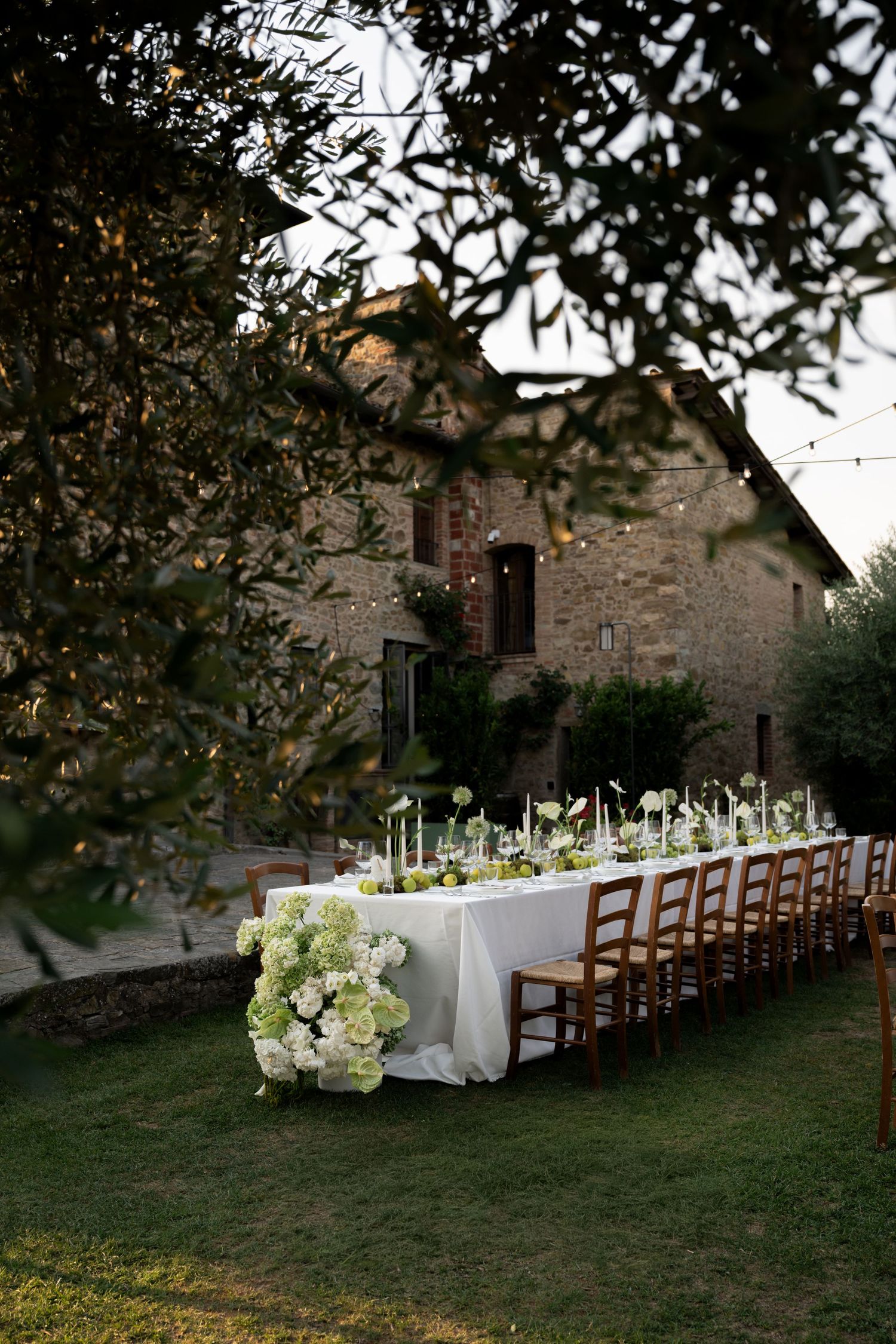Elegant outdoor wedding reception table with white floral arrangements set on green lawn before a rustic Tuscan stone villa.