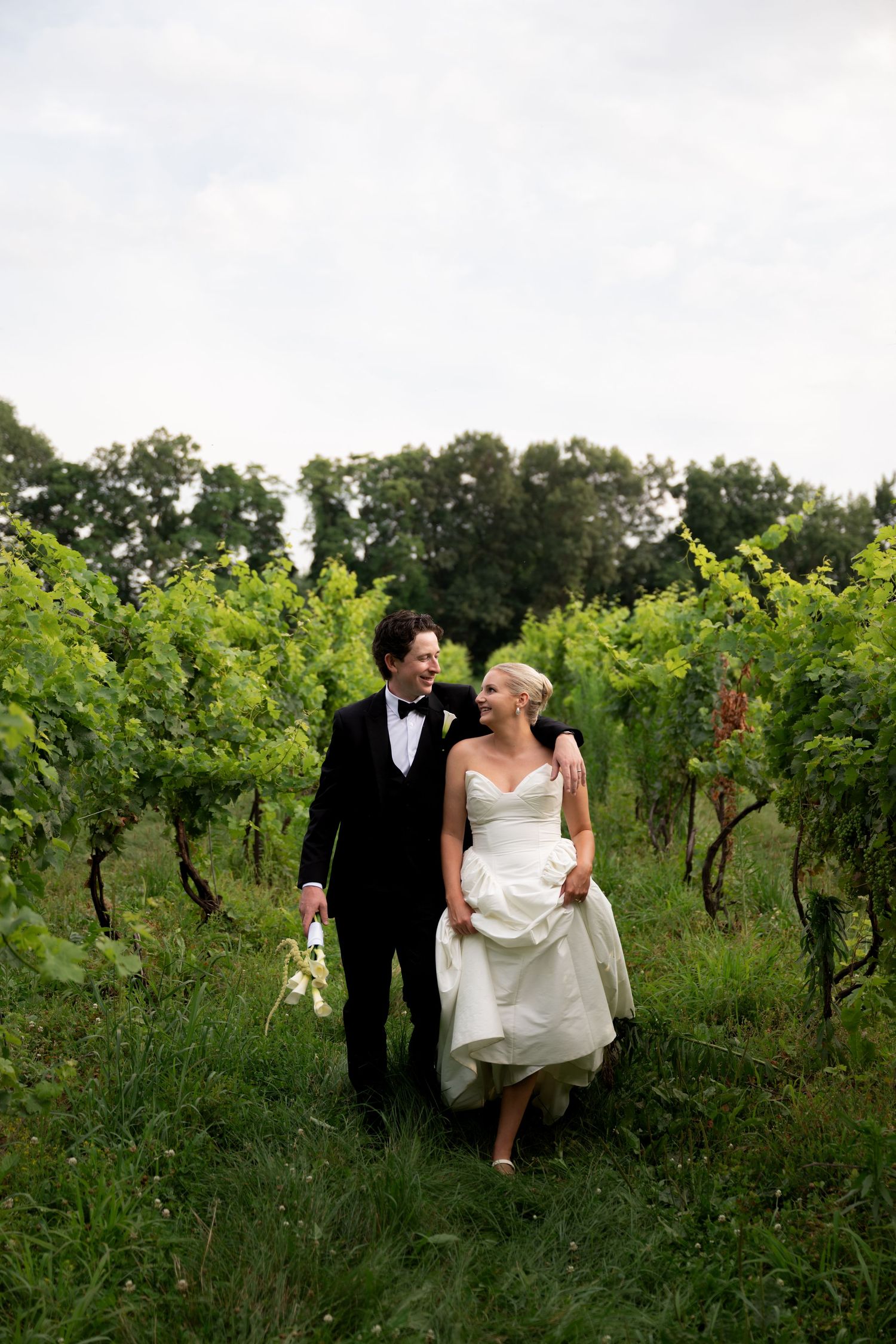 A bride and groom walk hand in hand through lush green vineyard rows on their wedding day.