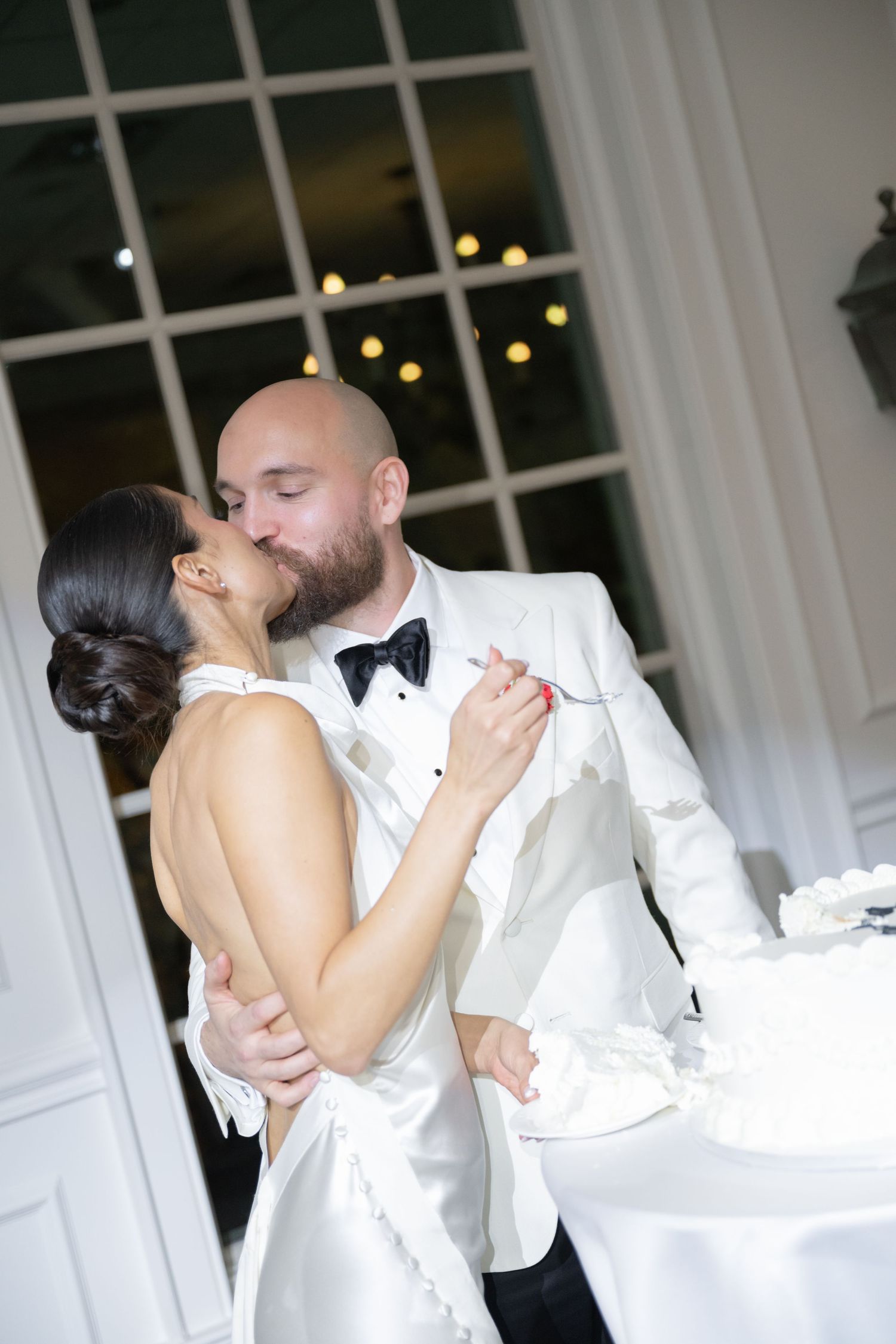 Romantic first dance moment between newlyweds in formal attire near a window with twinkling lights.