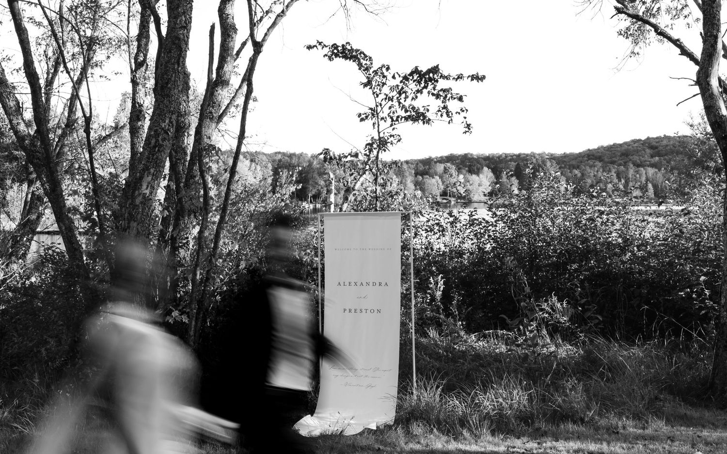 A blurred figure walks past a memorial stone marker in a black and white wooded landscape.