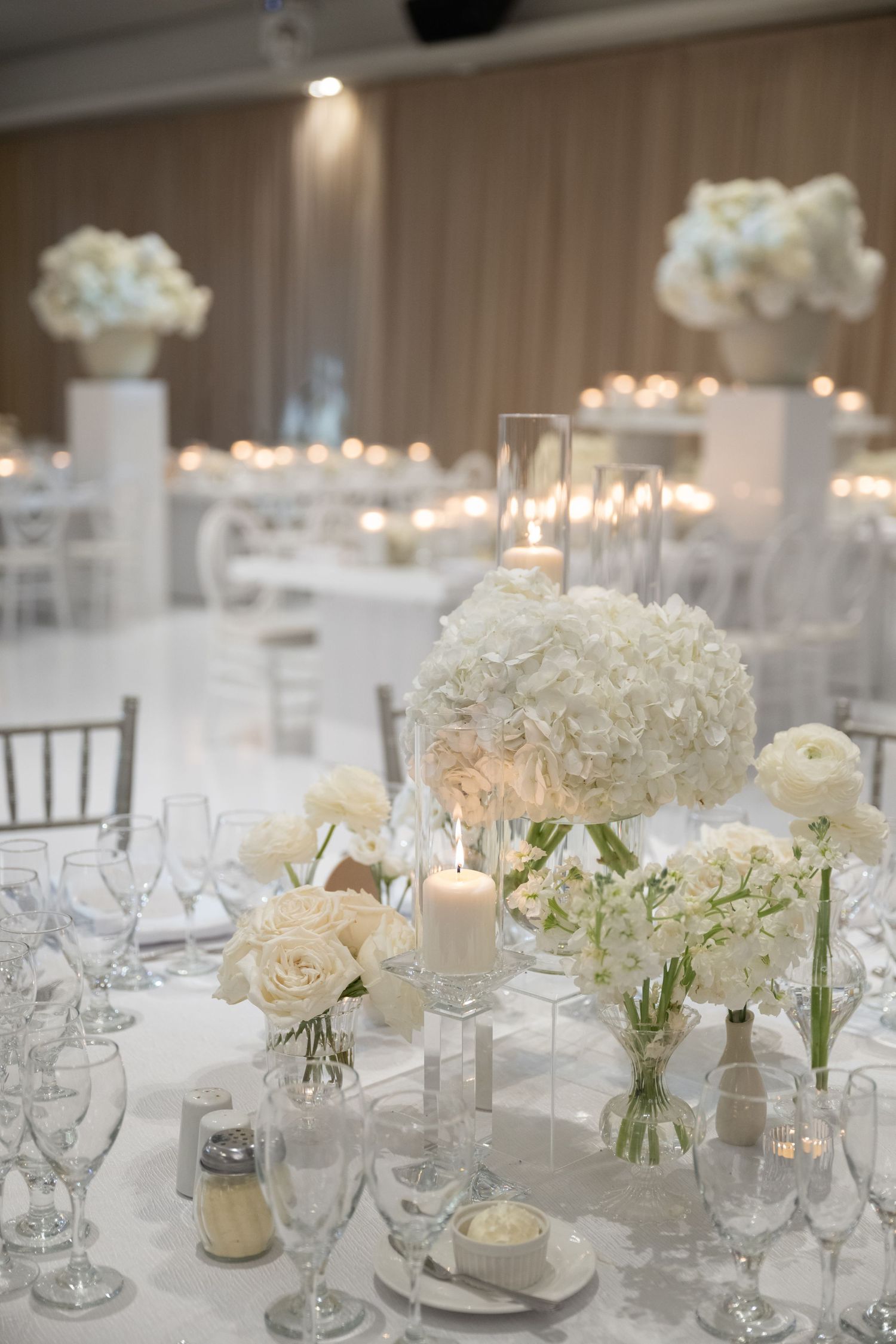 Elegant white wedding reception table with hydrangeas, roses, and candlelight centerpieces on a white tablecloth.