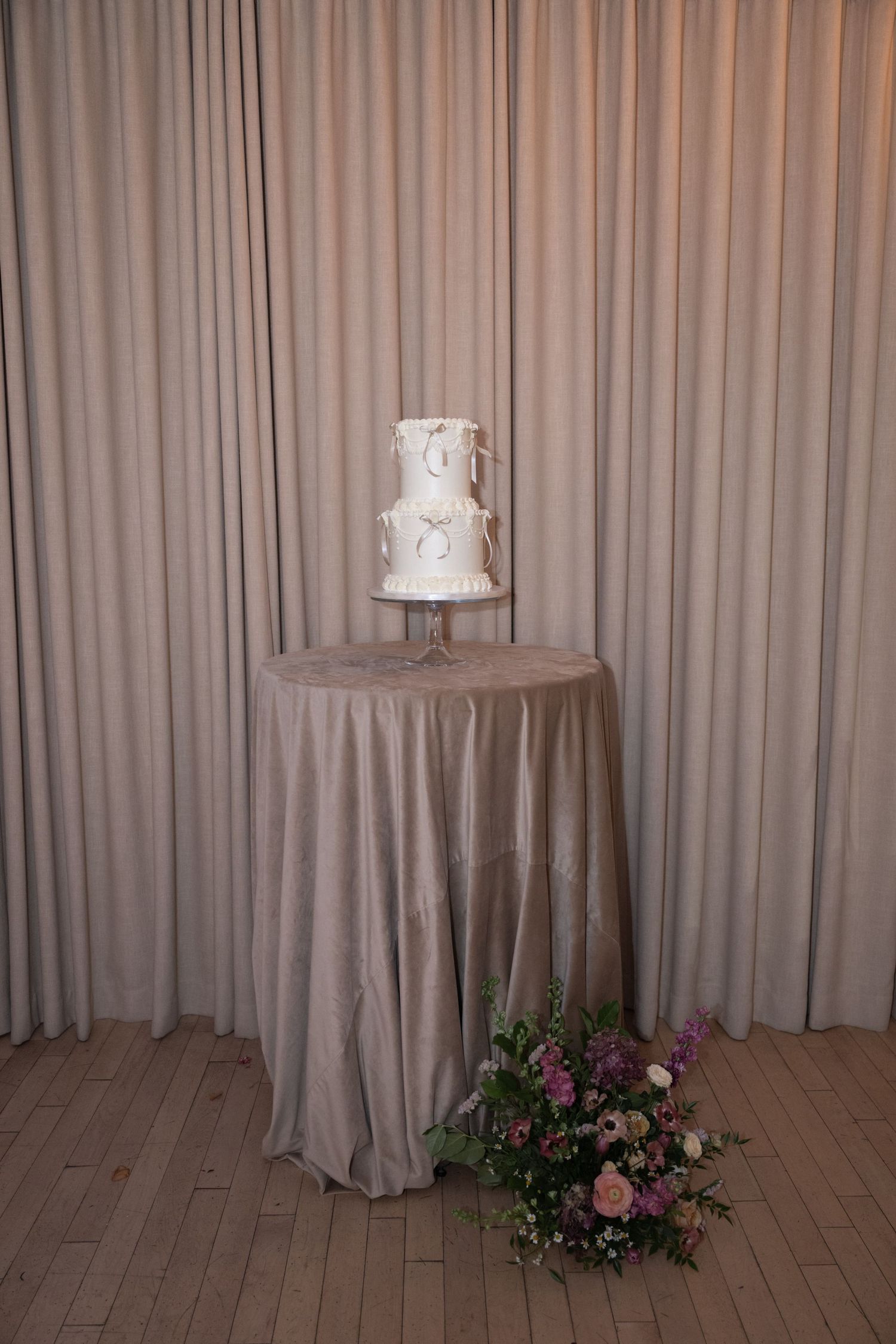 A tall white wedding cake displayed on a draped table with pink floral arrangements against a beige curtain backdrop.