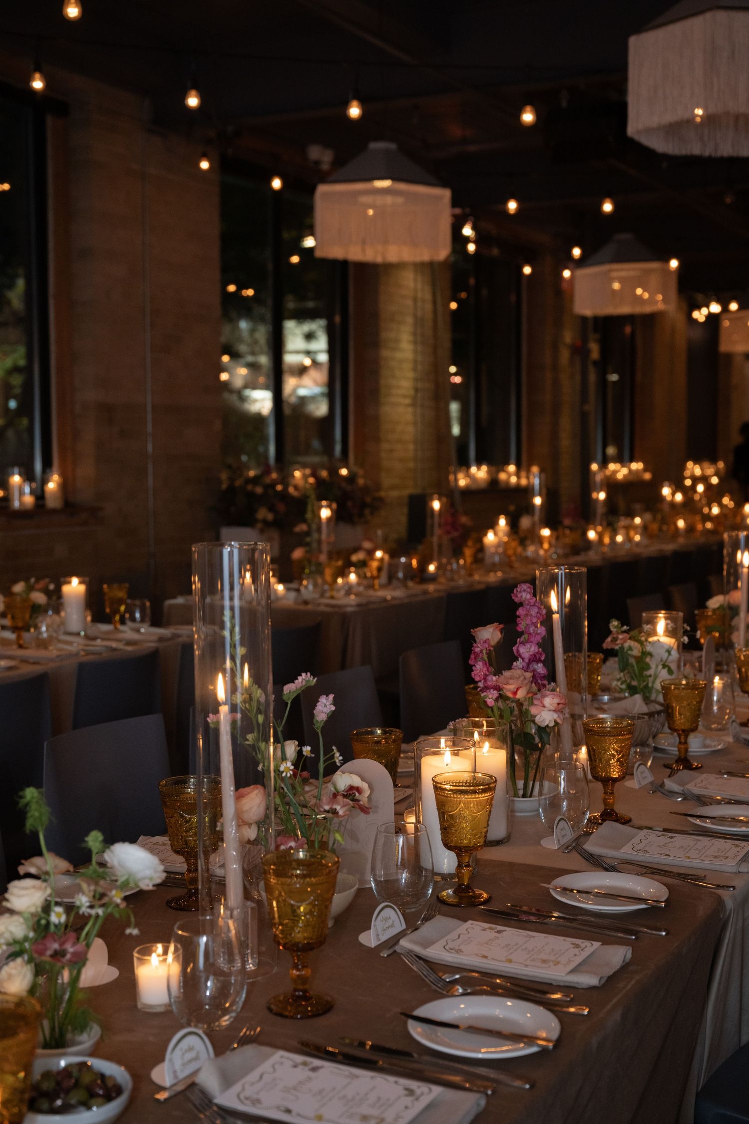 Elegant wedding reception table with candlelight, amber glassware and floral arrangements under warm string lighting and lanterns.