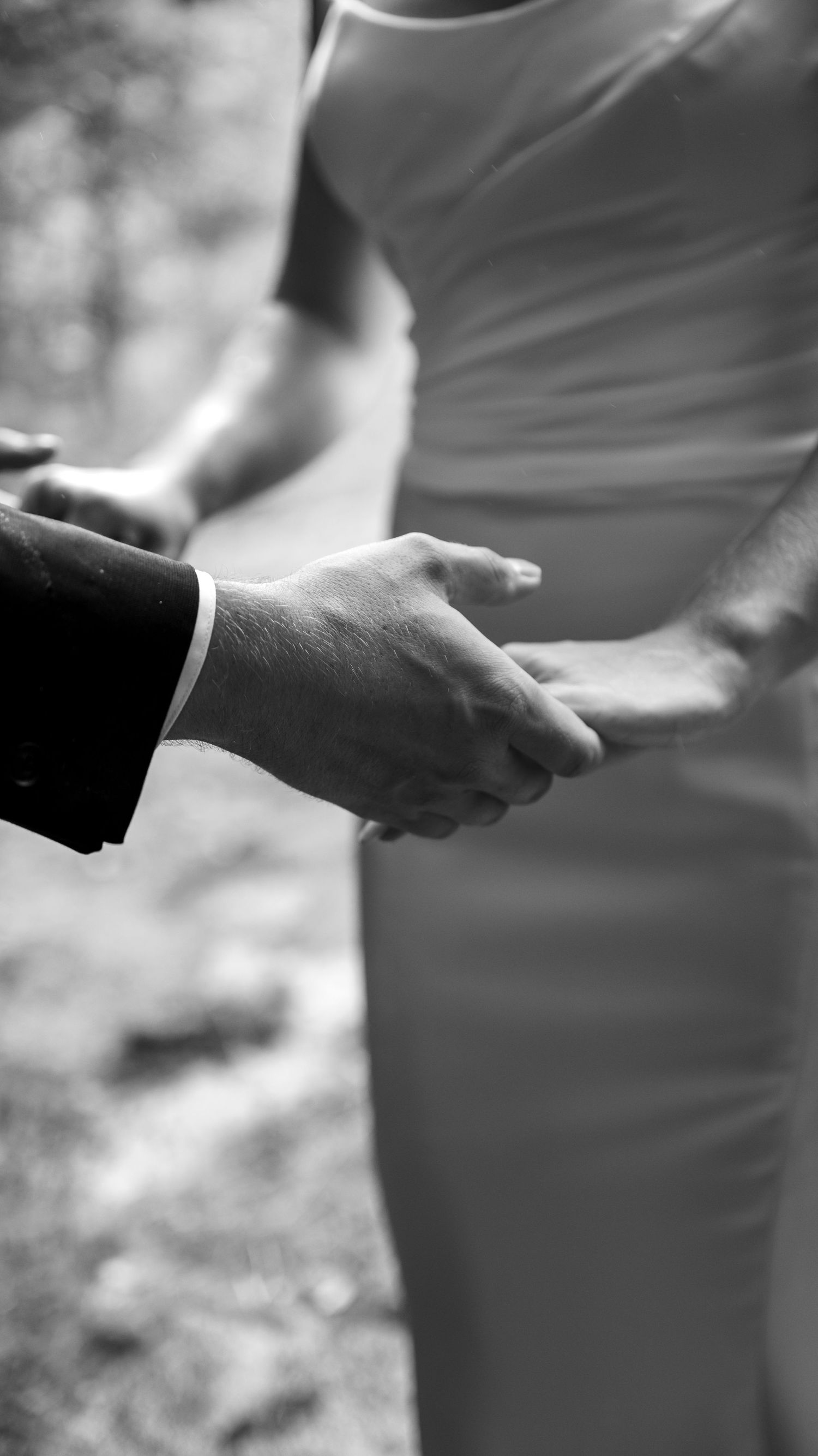 Close-up black and white photo of clasped hands during an intimate outdoor ceremony moment.