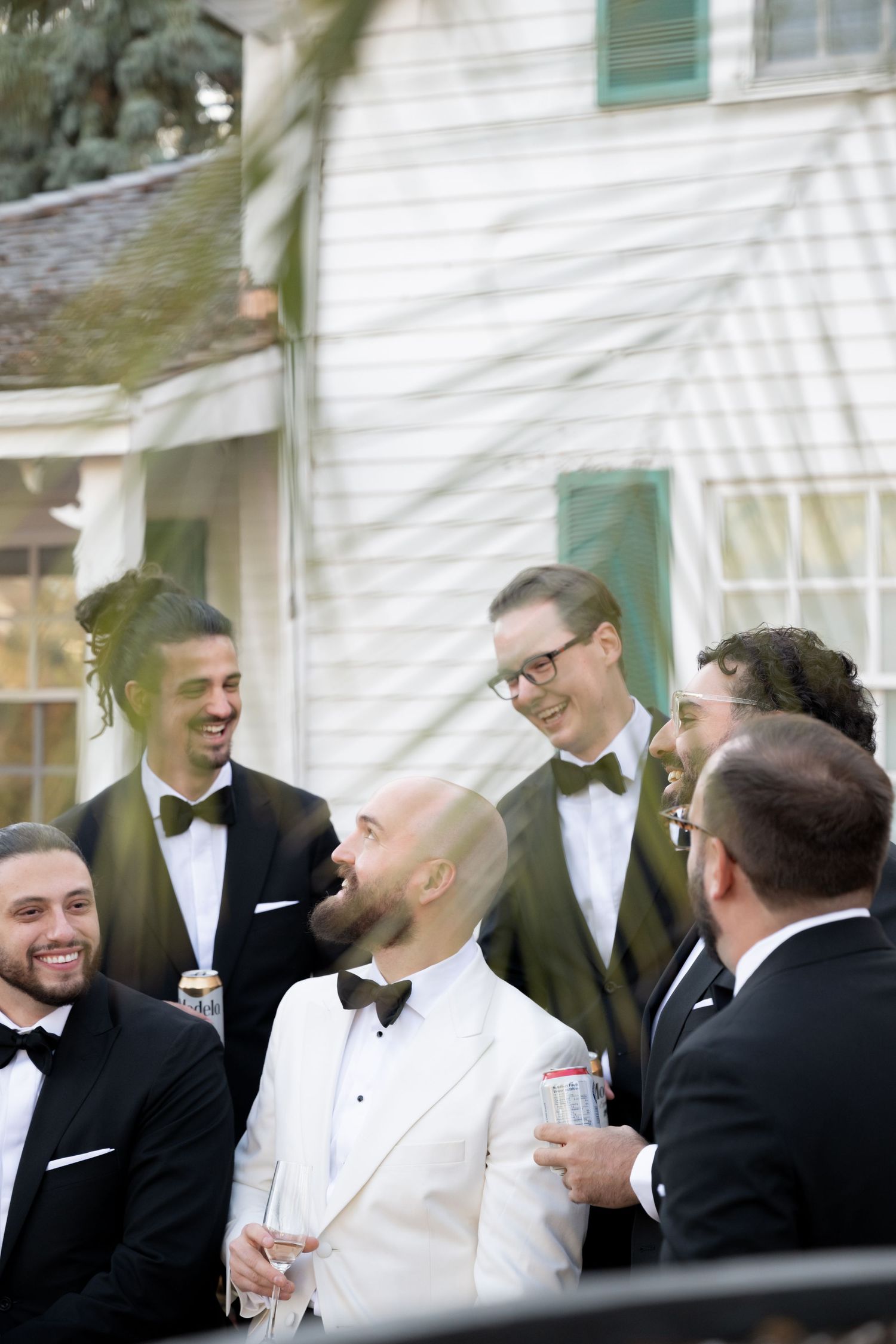 A group of men in formal black and white tuxedos share laughs and drinks at an elegant outdoor event.